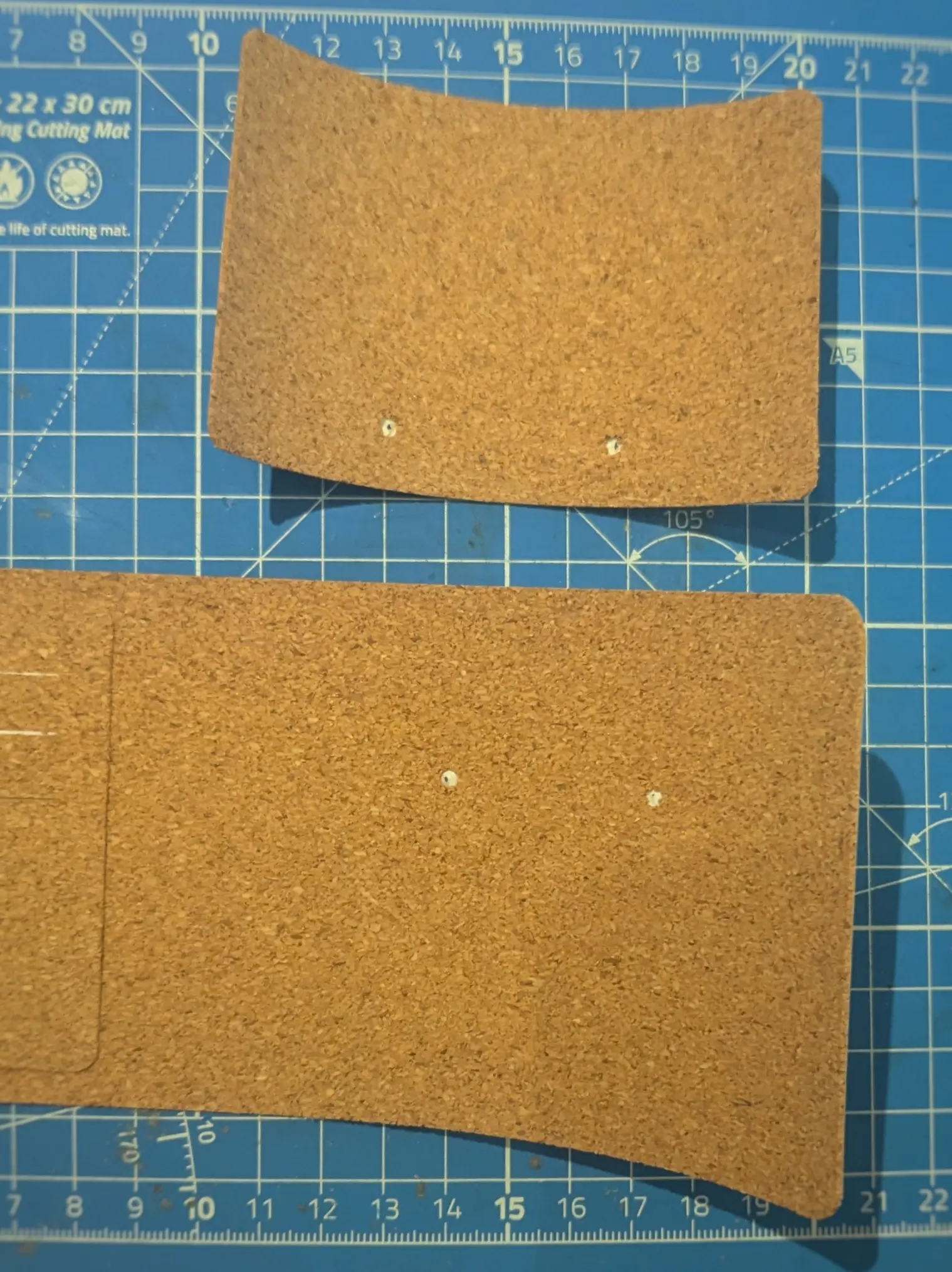 One smaller cork rectangle and the inner cork sleeve rectangle placed on a blue cutting mat.
Only the right-hand side of the inner sleeve is visible, showing that just like the smaller rectangle placed on top of it, it has two holes punched out.