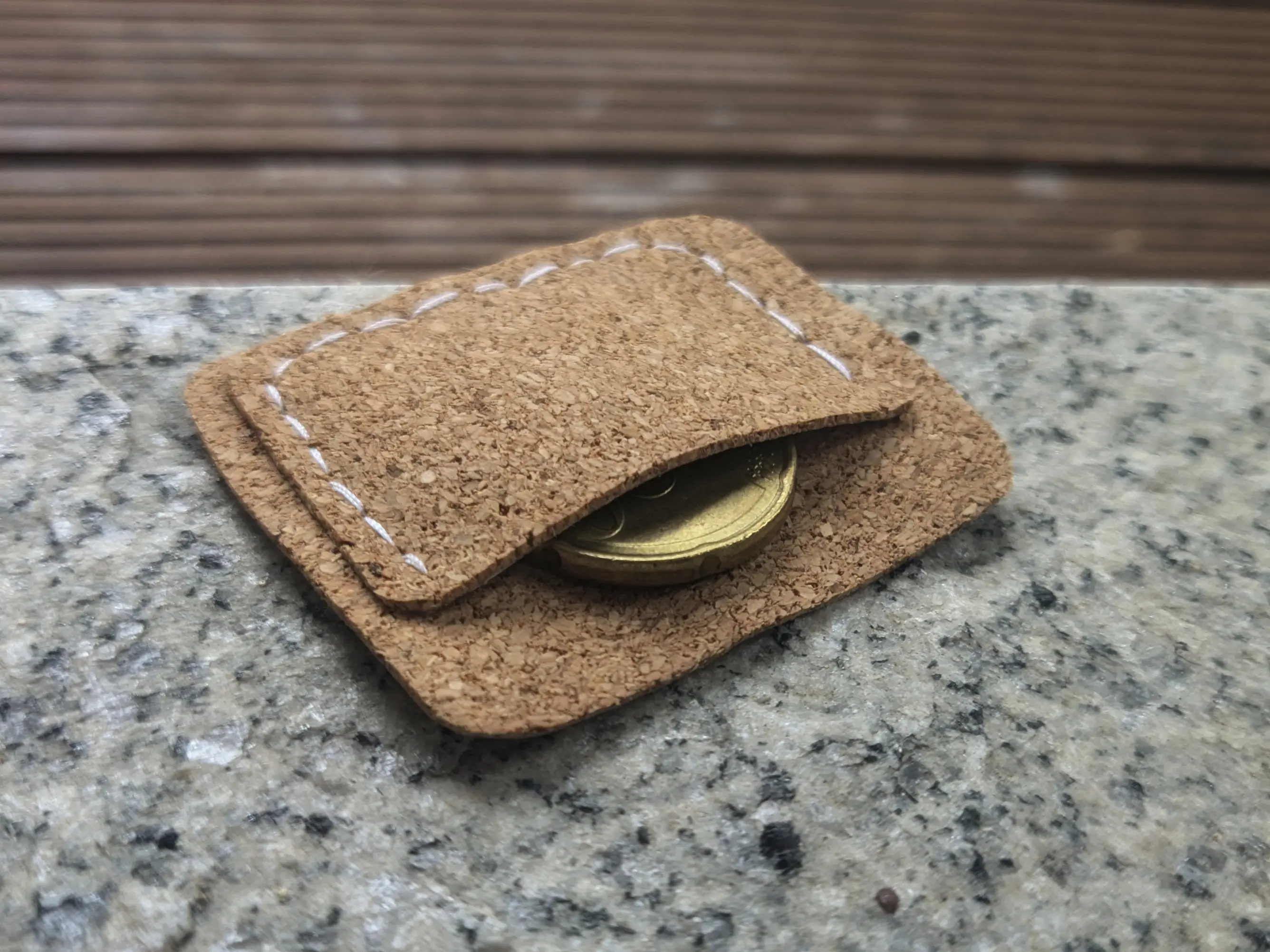 Small cork pocket placed on a stone surface. The piece itself is made out of two cork layers.
One slightly larger backing piece with rounded corners.
And a smaller front piece, which forms the pocket and has been glued onto the back roughly in the middle.
White stitches follow the front piece along the sides and bottom.
A coin has been placed in the pocket, showing off its functionality.