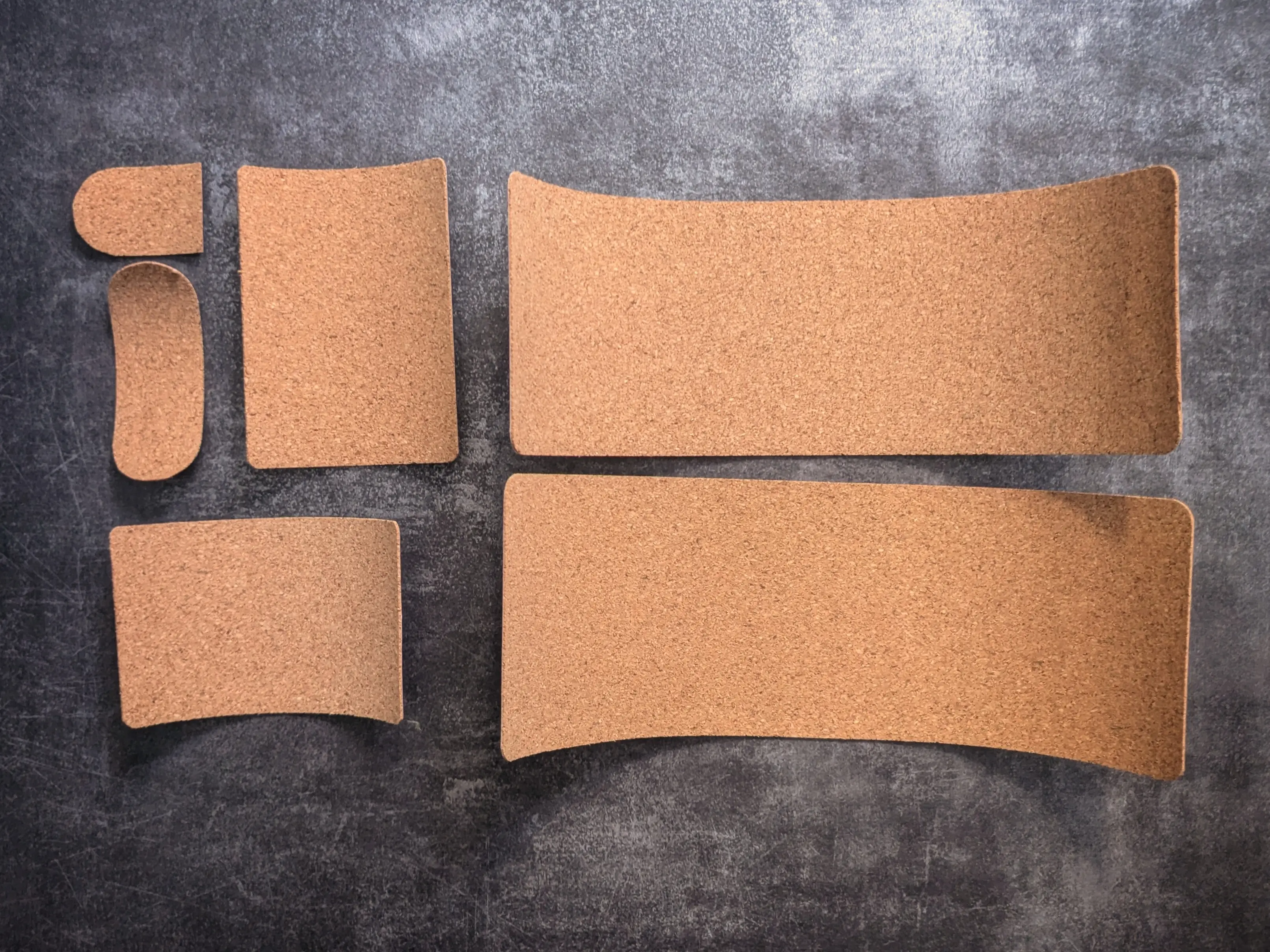 Six rectangular cork pieces of various sizes with rounded corners placed on a dark background.
Two wider pieces of the same size are placed on the right, while the other four are placed on the left.
All pieces curl upwards a little bit, still being slightly bent from the roll they used to be on.