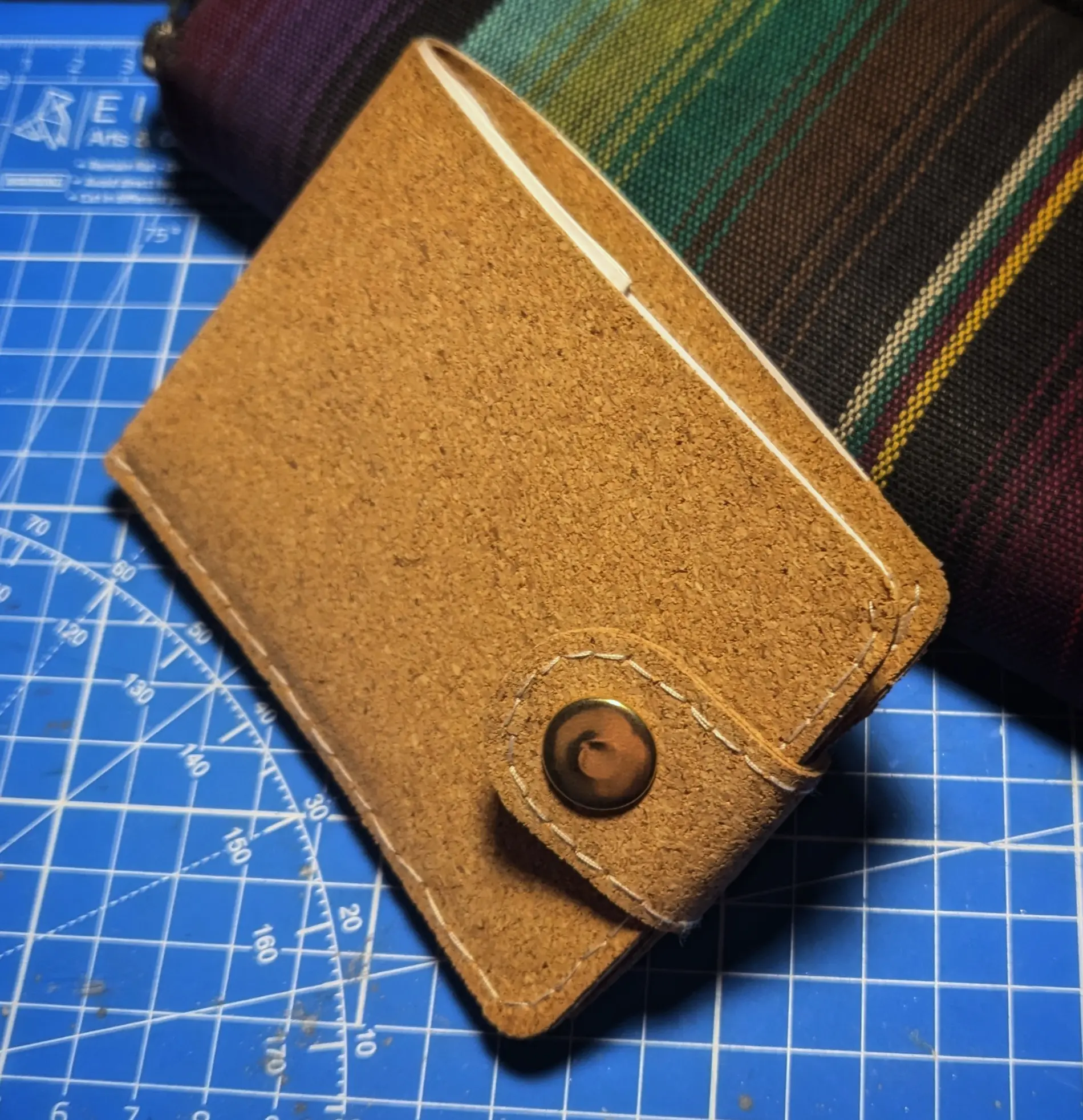 Closed cork wallet leaning against a longer wallet made from colorful fabric.
The cork wallet forms a U with a cork latch with a golden button forcing it shut.
Both the latch and the outer cork shell have white accent stitches running along their outer edge.
