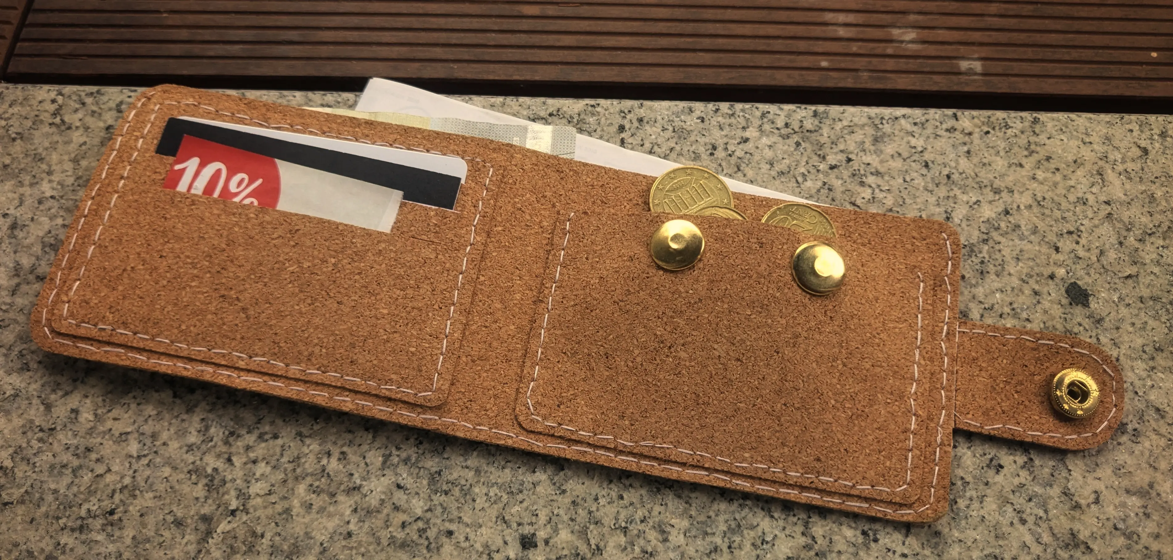 Opened cork wallet placed on a stone surface.
A banknote is sticking out of a longer pocket running along the entire length of the wallet.
On the inside of the wallet are two smaller rectangular pieces.
On the left-hand side of the inside is an ID card holder, which has three cards in total, one in every tier, sticking out slightly above their slots.
The pocket on the right-hand side is open on the top and has two buttons that can be used to close it. A couple of coins have been placed sticking out from it, indicating that it can be used to store coins.
White stitches are running along the outer edges of the wallet and the inside pockets.