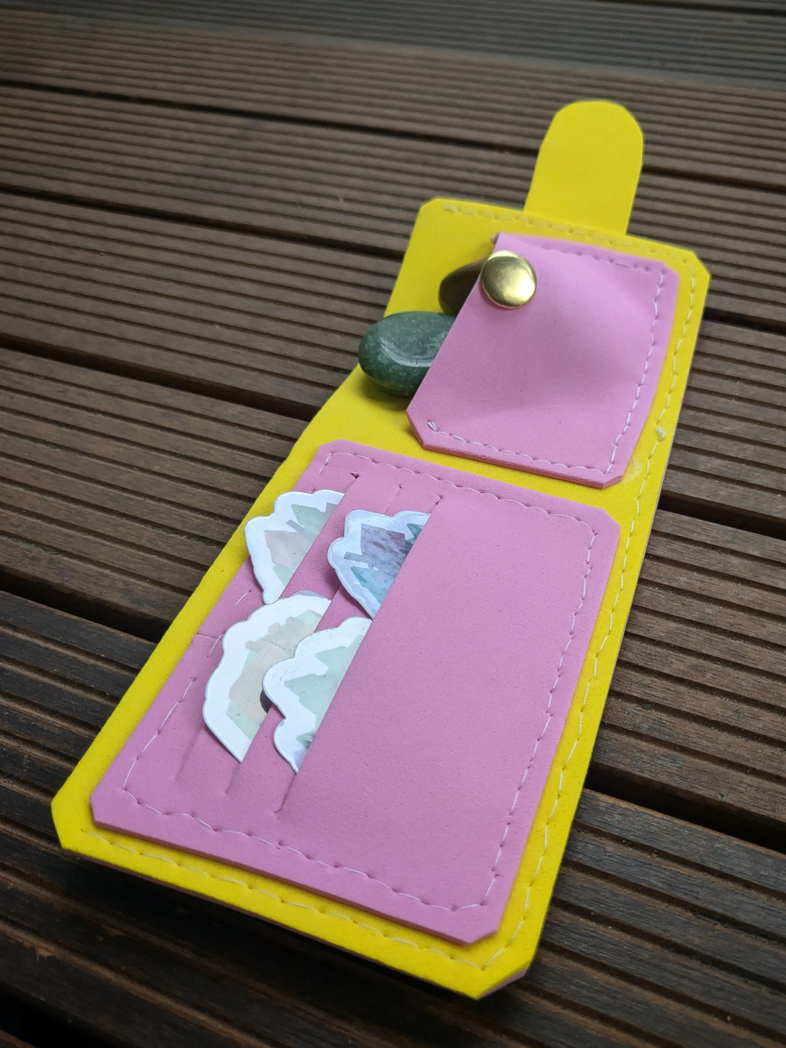 Long opened foam wallet placed on wooden boards.
The wallet is made from two colors: Yellow foam for the main sleeve material of the wallet: a rectangle with chamfered corners and a small latch extending from one side.
Two functional pieces, made from pink foam, have been sewn on to the inside of the yellow sleeve.
On the left half of the wallet is a rectangular piece with three slots in it, which have been filled with stickers, showing of the inside pockets.
On the right half of the wallet is a shorter rectangular piece with a button on it. It has been filled with two small stones, indicating that it is a larger pocket.