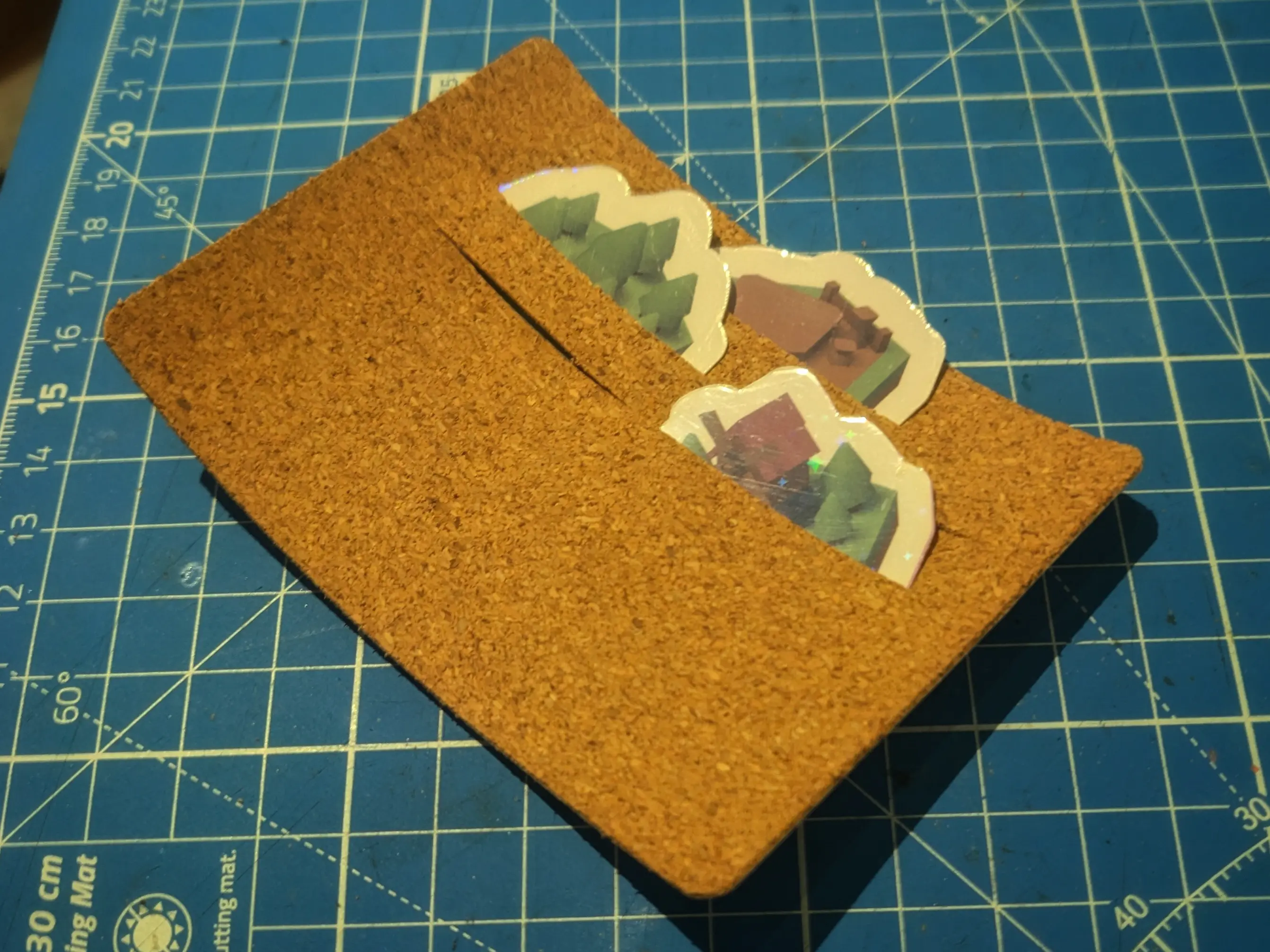 Rectangular cork piece placed on a blue cutting mat.
It has three slots cut into the upper half that span almost the entire width. Each slot has a piece of paper sticking out of it to show where they are.