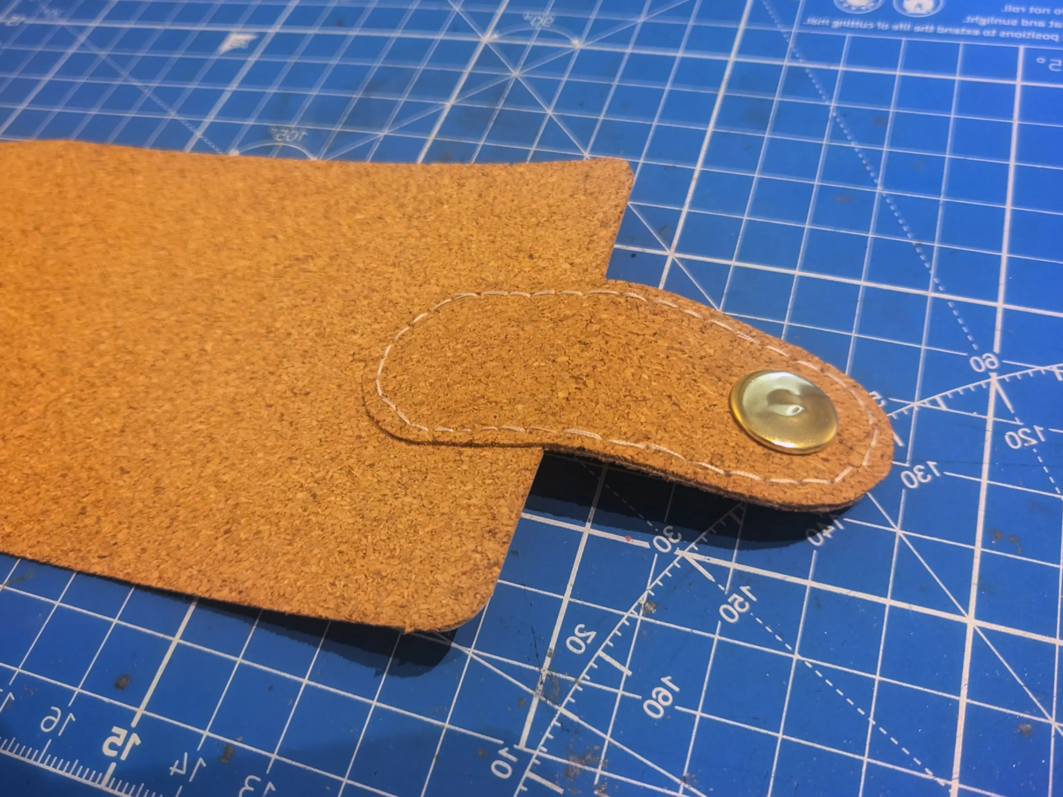 The latch piece glued to the outer sleeve (one of the two large cork rectangles). It is centered along the short side and overlapping it by one third.
A white outline has been stitched on along the outer edge.