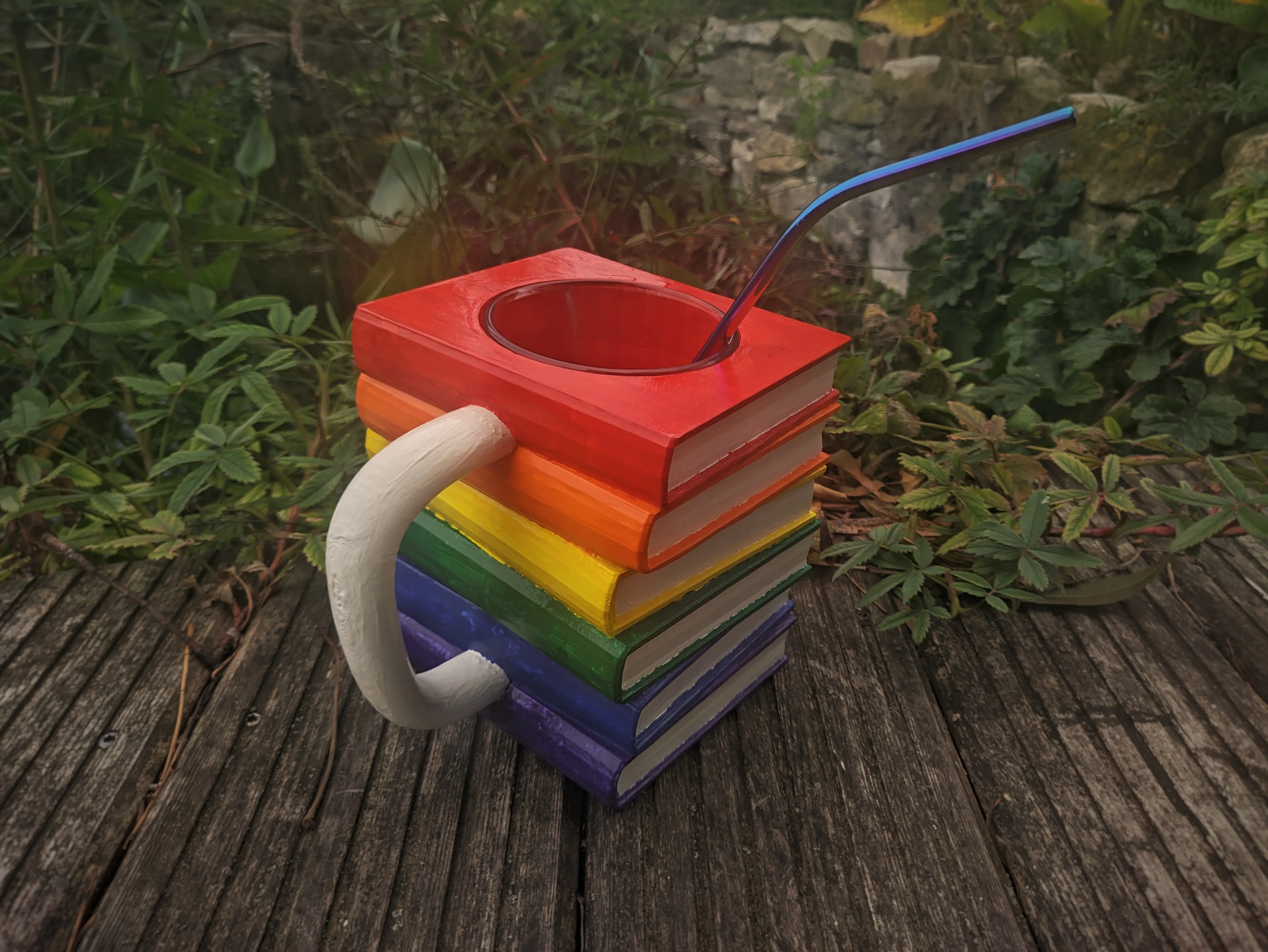 The painted mug with a glass and straw inside placed on wooden planks.
Every book has been painted a single color, forming the colors of the pride flag in the correct order when stacked.
The handle and pages have been painted an off-white color.