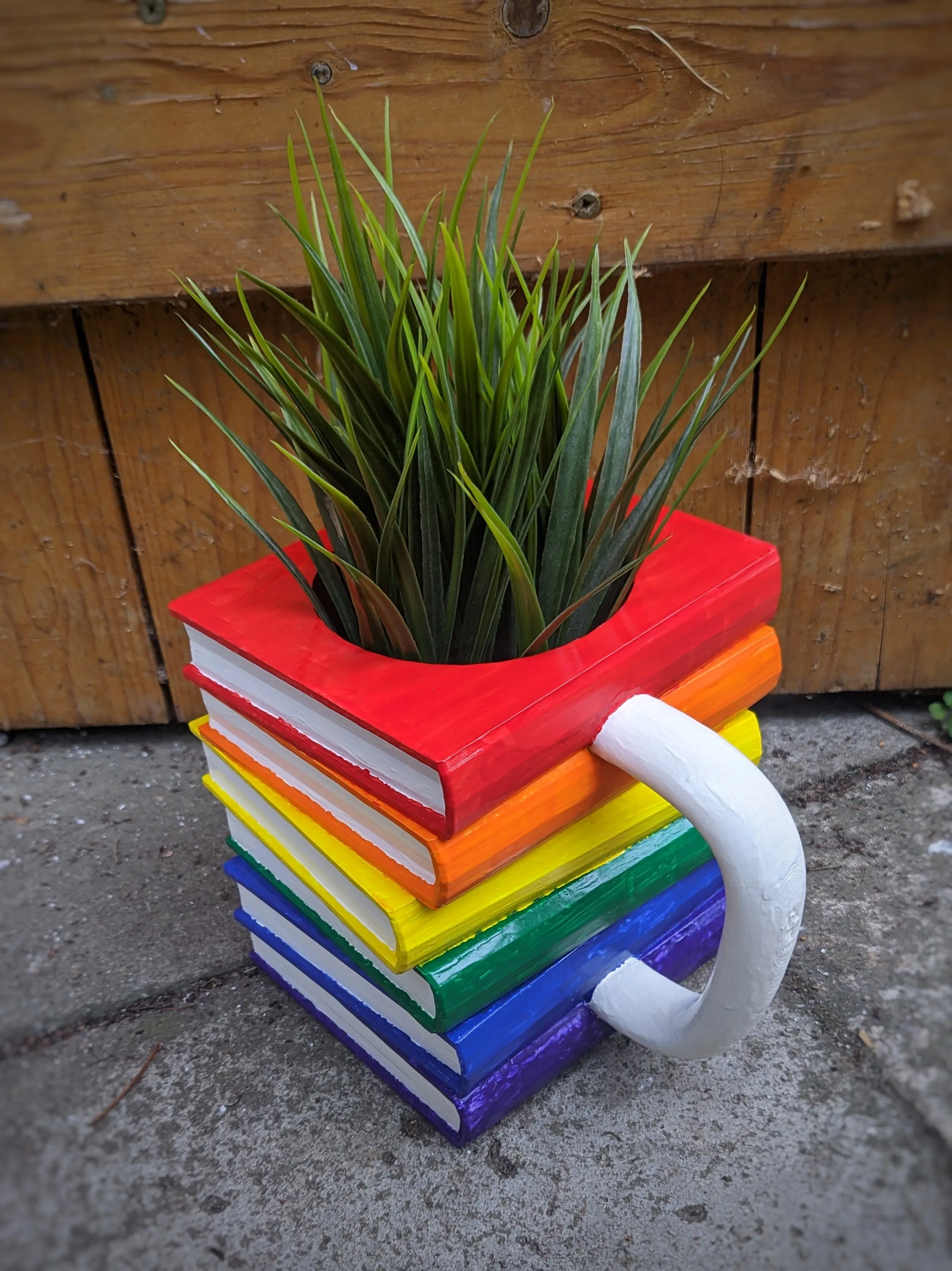 The painted mug has been placed on stone floor tiles in front of a wooden door.
Instead of the glass, a plant has been placed inside the cutout hole.
The plant has thin and long leaves, almost making it look like hair is growing from it.