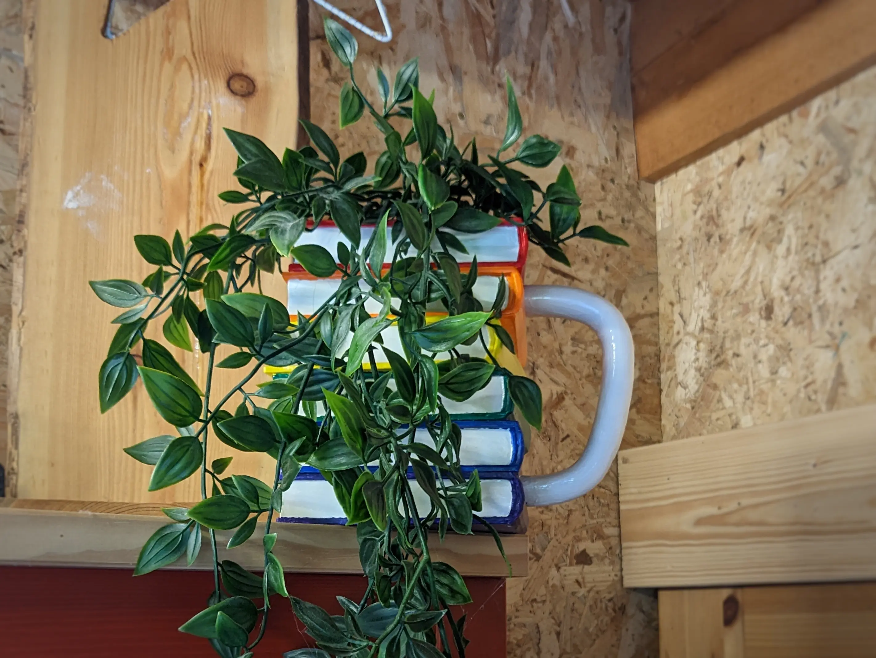 The same mug has been placed on a shelf with a climbing plant inside.
The plant hangs down over the books and hides most of them, except for some rainbow colours shining through.