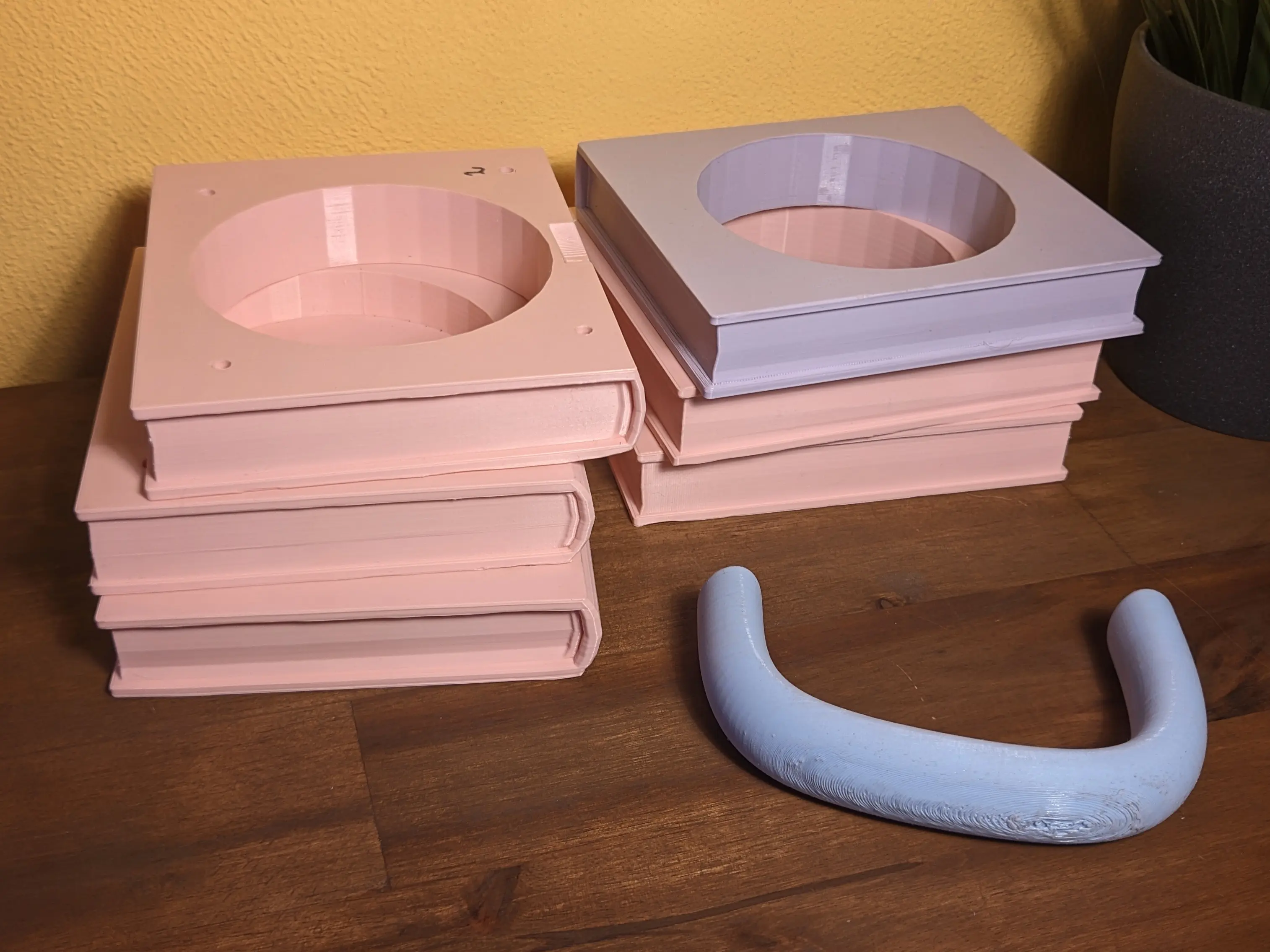 Two stacks of 3D-printed books with a hole in the middle placed on a wooden work surface.
The stacks have three books each, stacked without order. In front of the stacks lies the 3D-printed handle.
The parts have different colors.