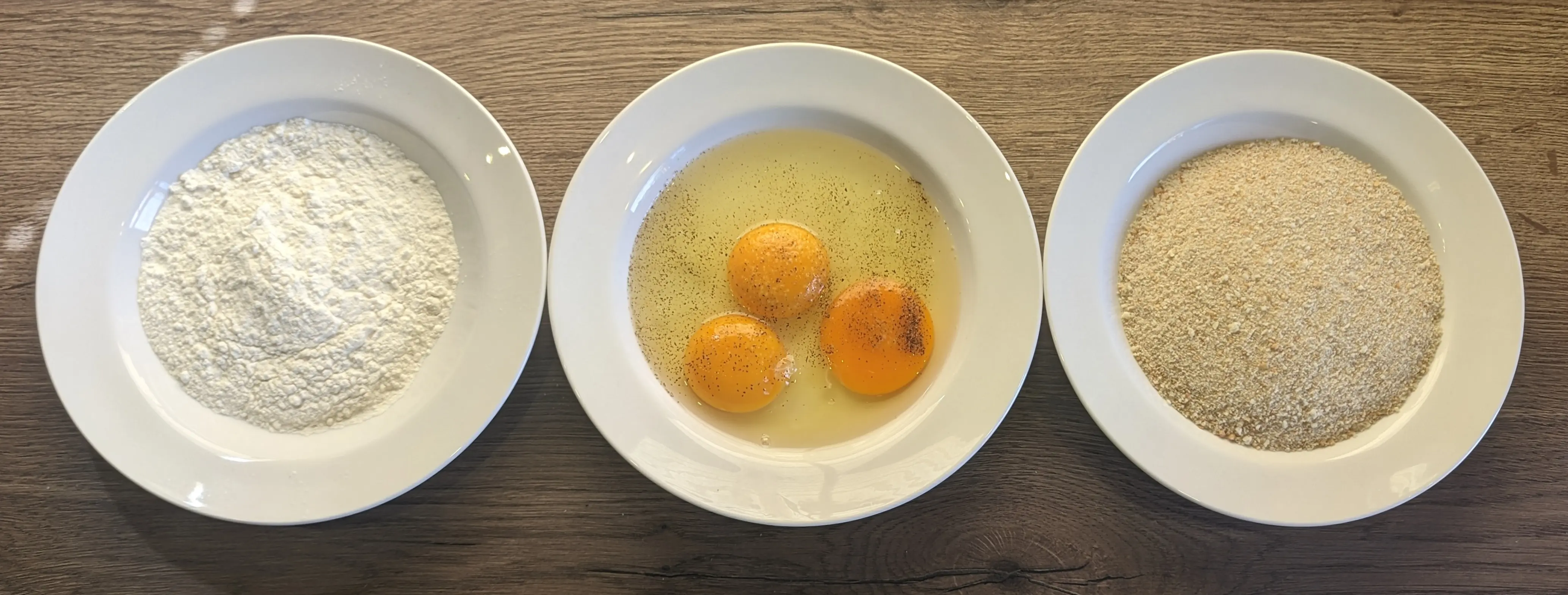 Three plates are placed in a line on a work surface.
The leftmost plate is filled with flour.
The center plate contains three unbeaten eggs as well as some salt and pepper.
The plate on the right is filled with breadcrumbs.