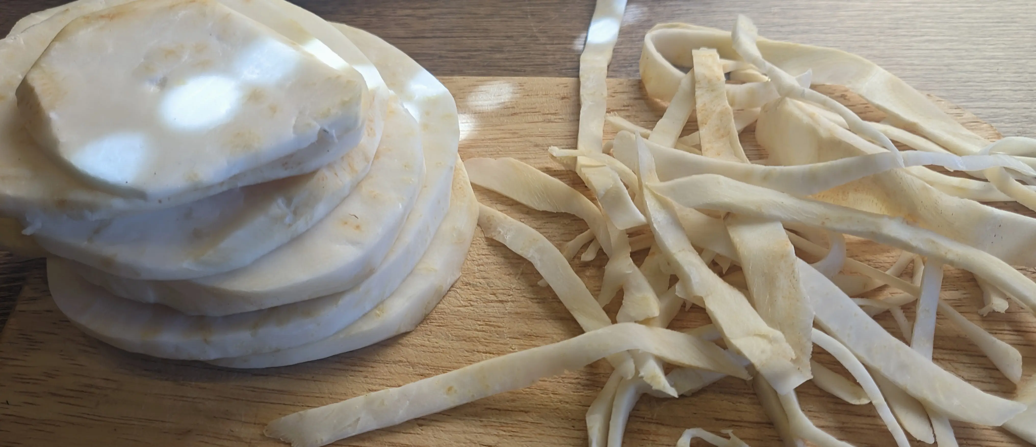 A stack of celeriac slices with a straight edge on a cutting board. Next to it is a stack of celeriac shavings, left from peeling the edge.