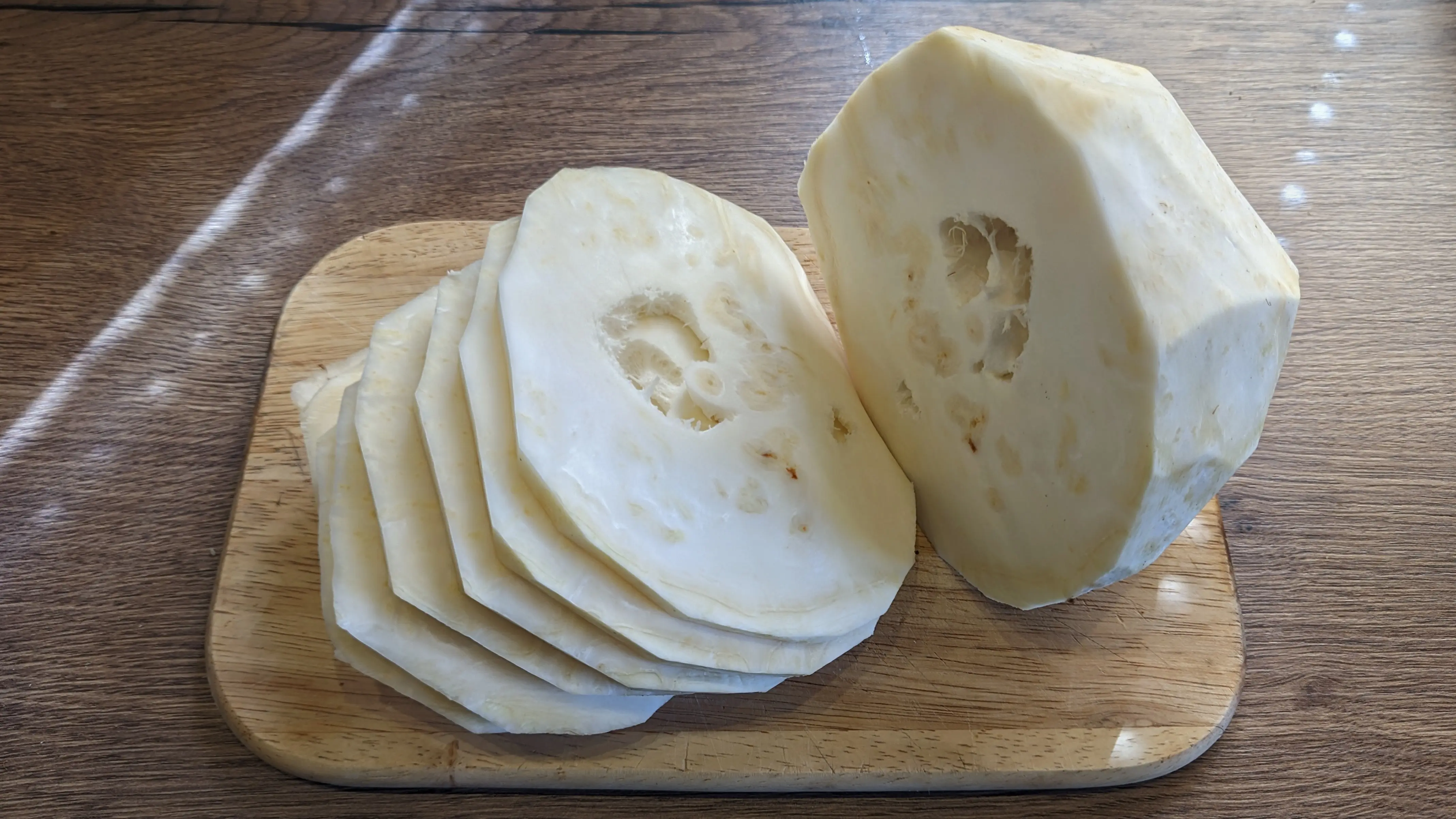 White peeled celeriac on a small cutting board.
Half of the celeriac has already been cut into slices, which are stacked on the left.
You can make out larger holes in the center of the larger slices and the untouched half.
