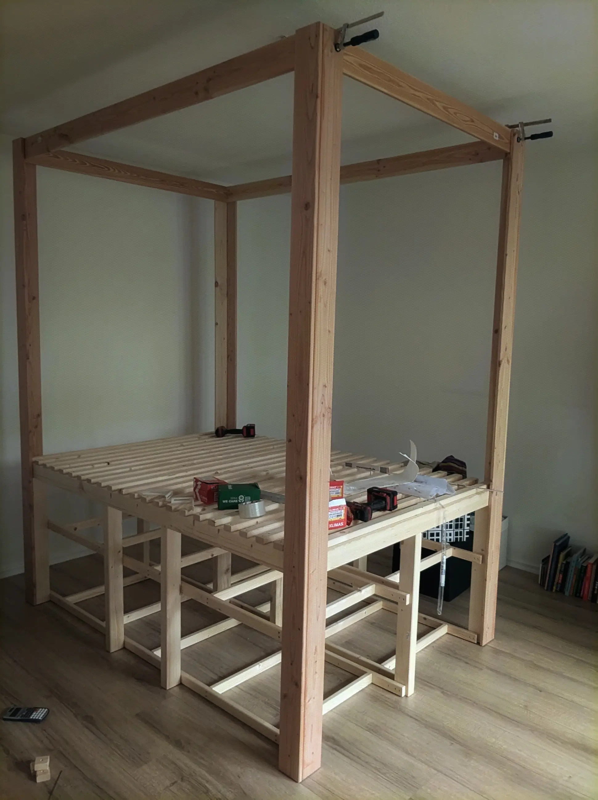 The same wooden structure, again, but from farther away.
The bed slats have been fully assembled, and long wooden beams have been mounted on the corners of the bed (the canopy legs).
On the top, the same type of beam is used to connect them together forming the canopy structure.
Two clamps are still holding on the front most beam The same wooden structure, again, but from farther away.
The bed slats have been fully assembled, and long wooden beams have been mounted on the corners of the bed (the canopy legs).
On the top, the same type of beam is used to connect them together forming the canopy structure.
Two clamps are still holding on the front most beam