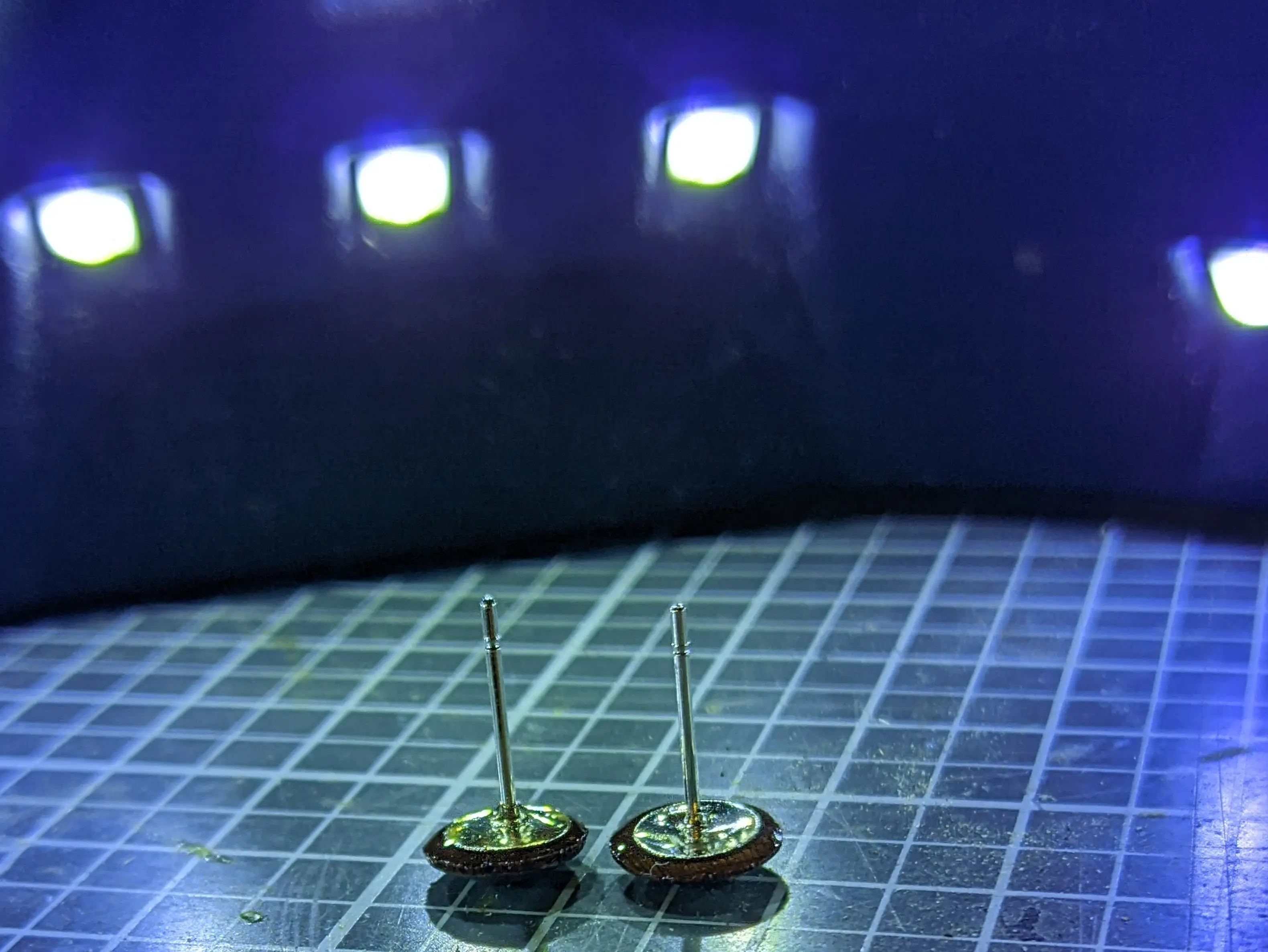 Earrings placed on a cutting mat. In the background some of the UV lamp LEDs can be made out.
The dome section is facing down. Metal rods have been attached to the back, with a visible blob of resin around the backing plates.