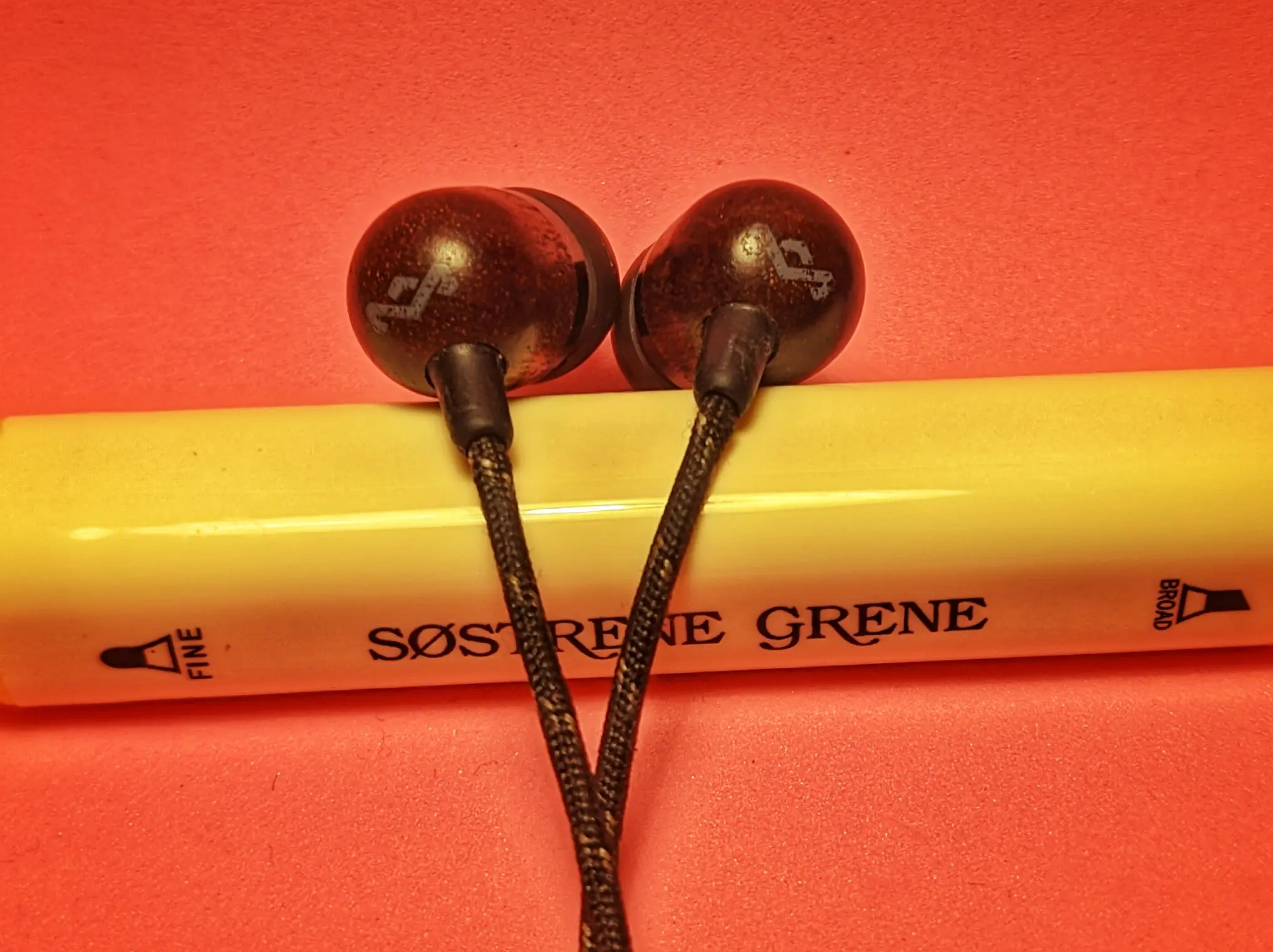 In-ear headphone earpieces hung over a pen. Two wires are running from the headphones out of the bottom of the screen. The headphone shell is made from a dark brown wood. The wood is rounded off at one end with the House Of Marley logo on it.