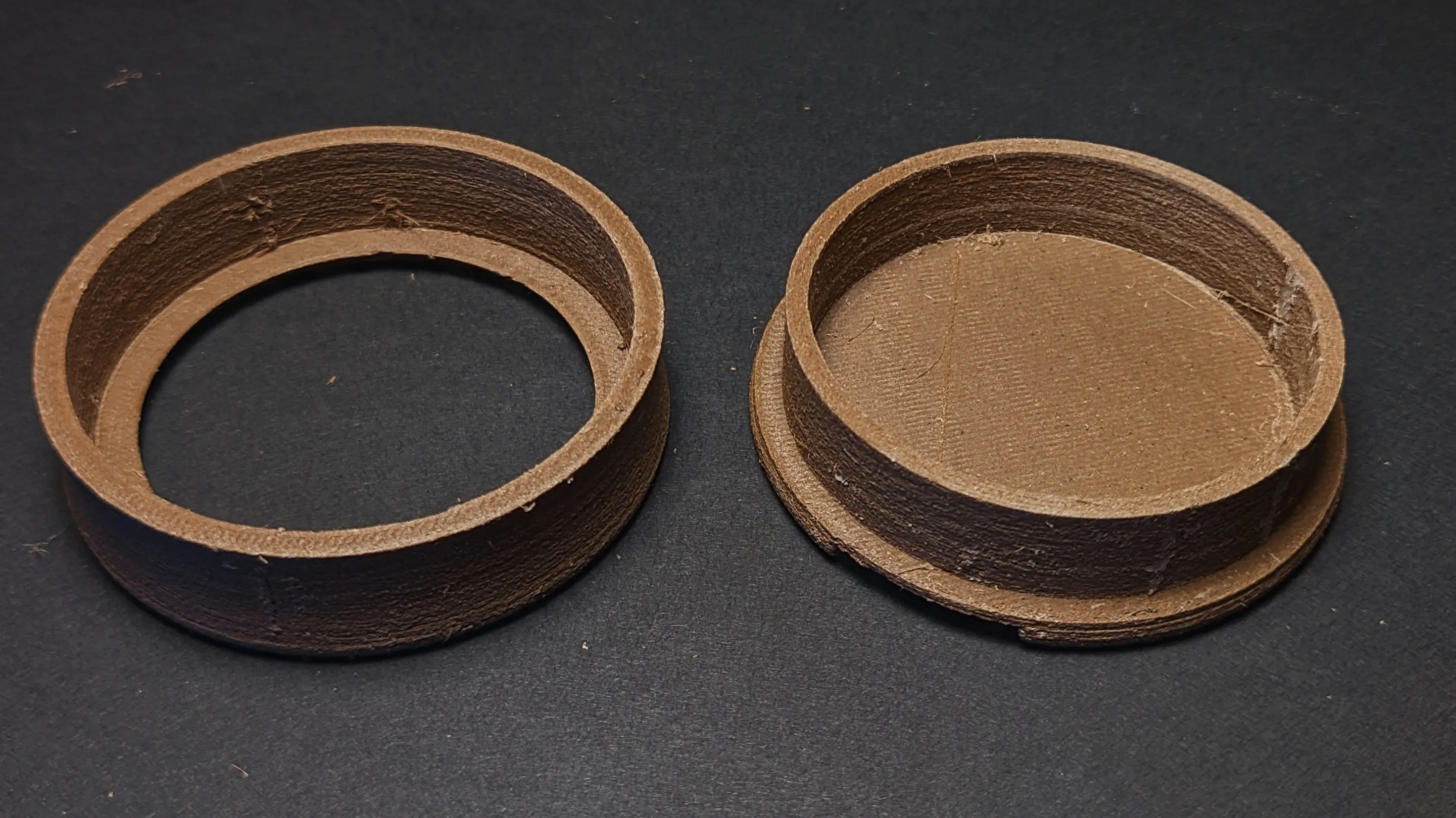 Printed top and bottom parts on a black backdrop.
They have been printed in a brown wood filament, which gives them a matte and slightly bumpy texture.
The bottom part is shaped like a hollow cylinder with an extended ledge at the bottom, closing it off, and a small rectangular cutout on one side.
The top part is an open cylinder with a slight ledge on the top. It has been flipped upside down so that its top is facing down.