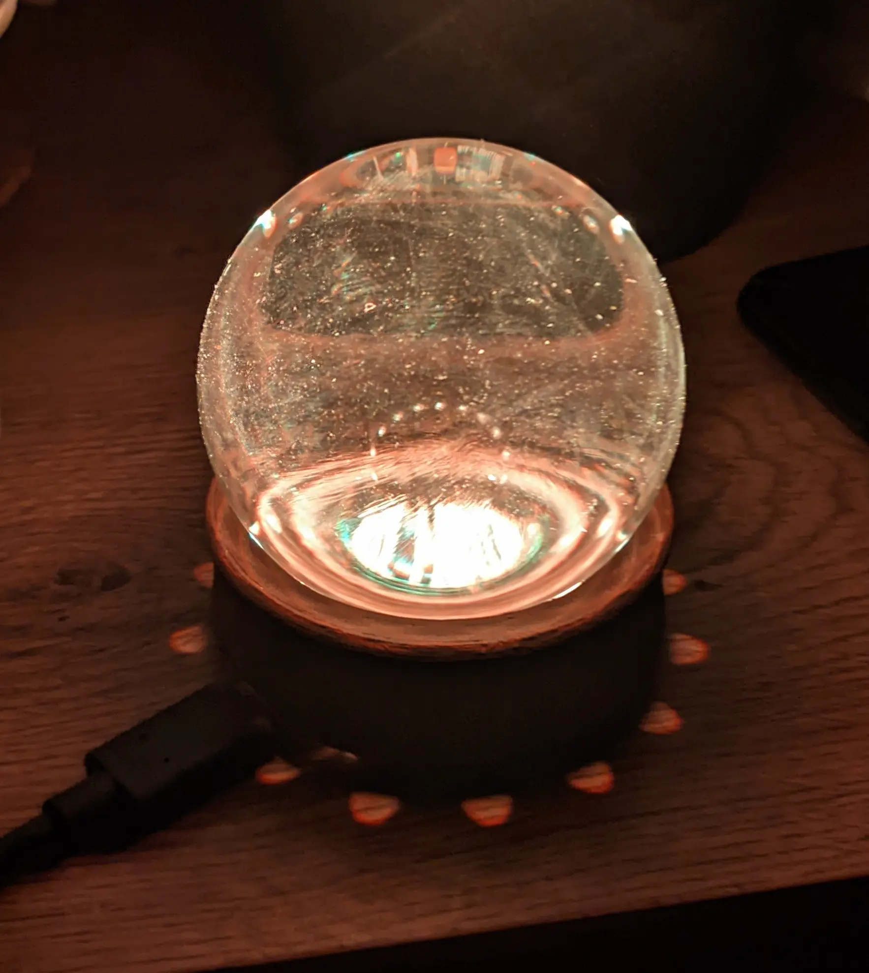GlowOrb placed on a nightstand besides a potted plant and a phone.
The lights inside partially reflect onto the night stand surface right next to the base.
Some fingerprints can be seen on the glass surface.
The crystal ball itself appears to glow white with a slight hint of orange.