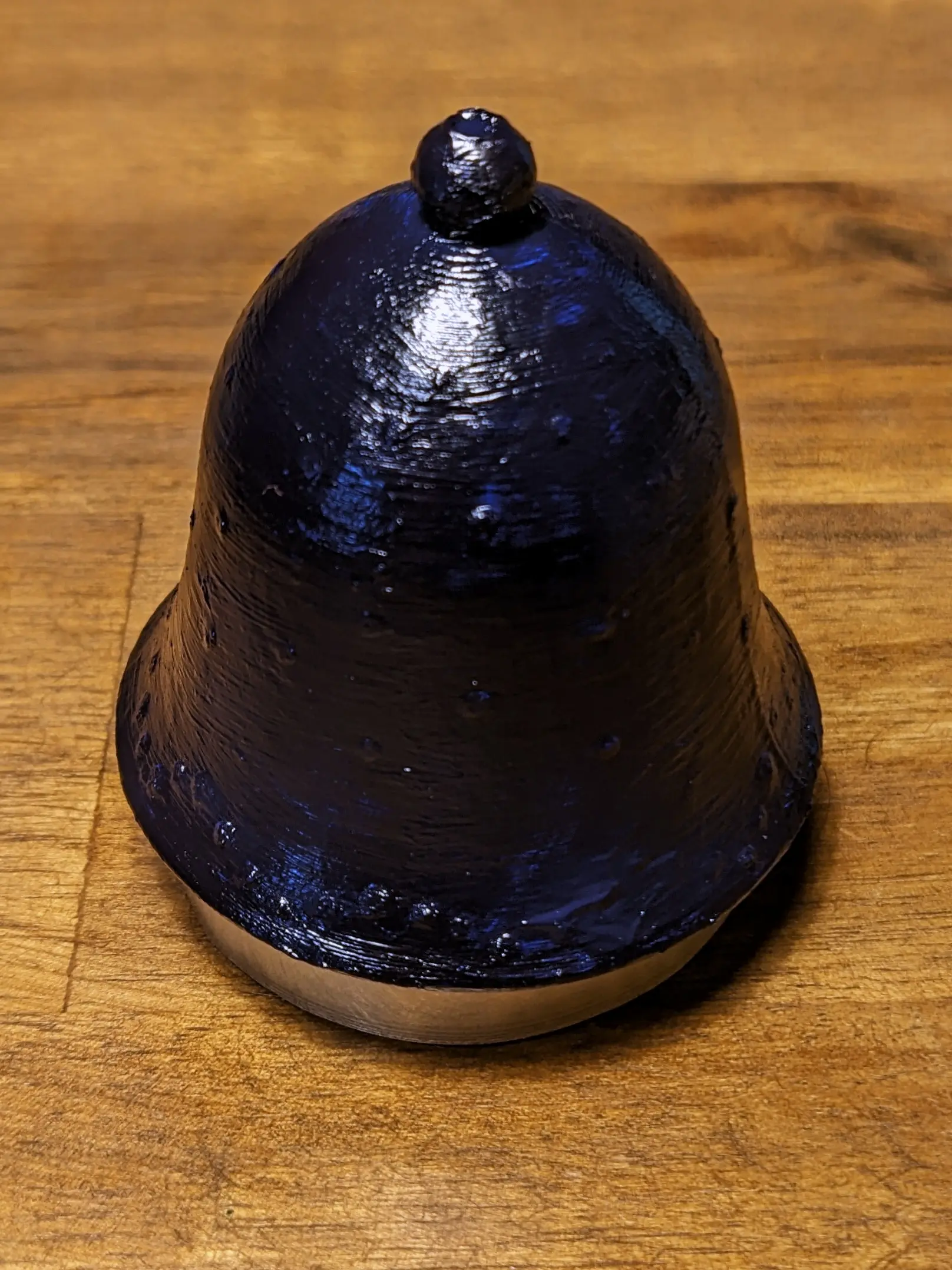 The bell has been placed on a small roll of tape, used to lift it off the table surface. The outside has been painted a dark blue, with some lighter spots shining through. Against the light, some layer lines are still visible, but especially in areas with little curvature, the layers of paint completely hide them. The bumps formed by the glue are still mostly visible, raising the blue paint slightly.