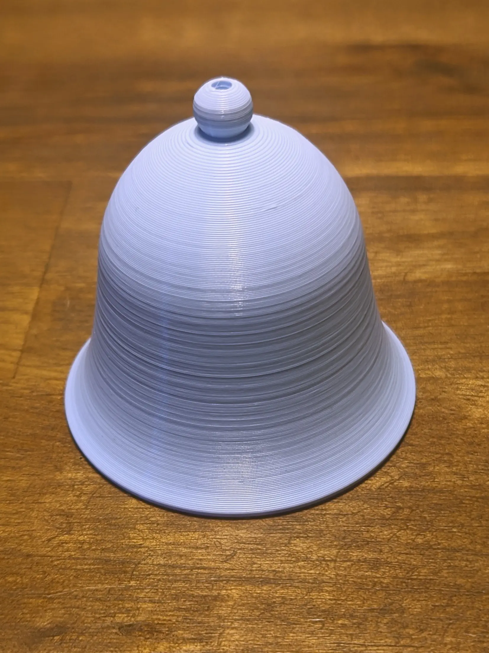 Light blue 3D-printed bell placed on a wooden desk. The lower part of the bell is flaring outwards. On the top of the bell a spherical accent piece with a hole in it is forming the bells crown. The layer lines are still very visible after printing.