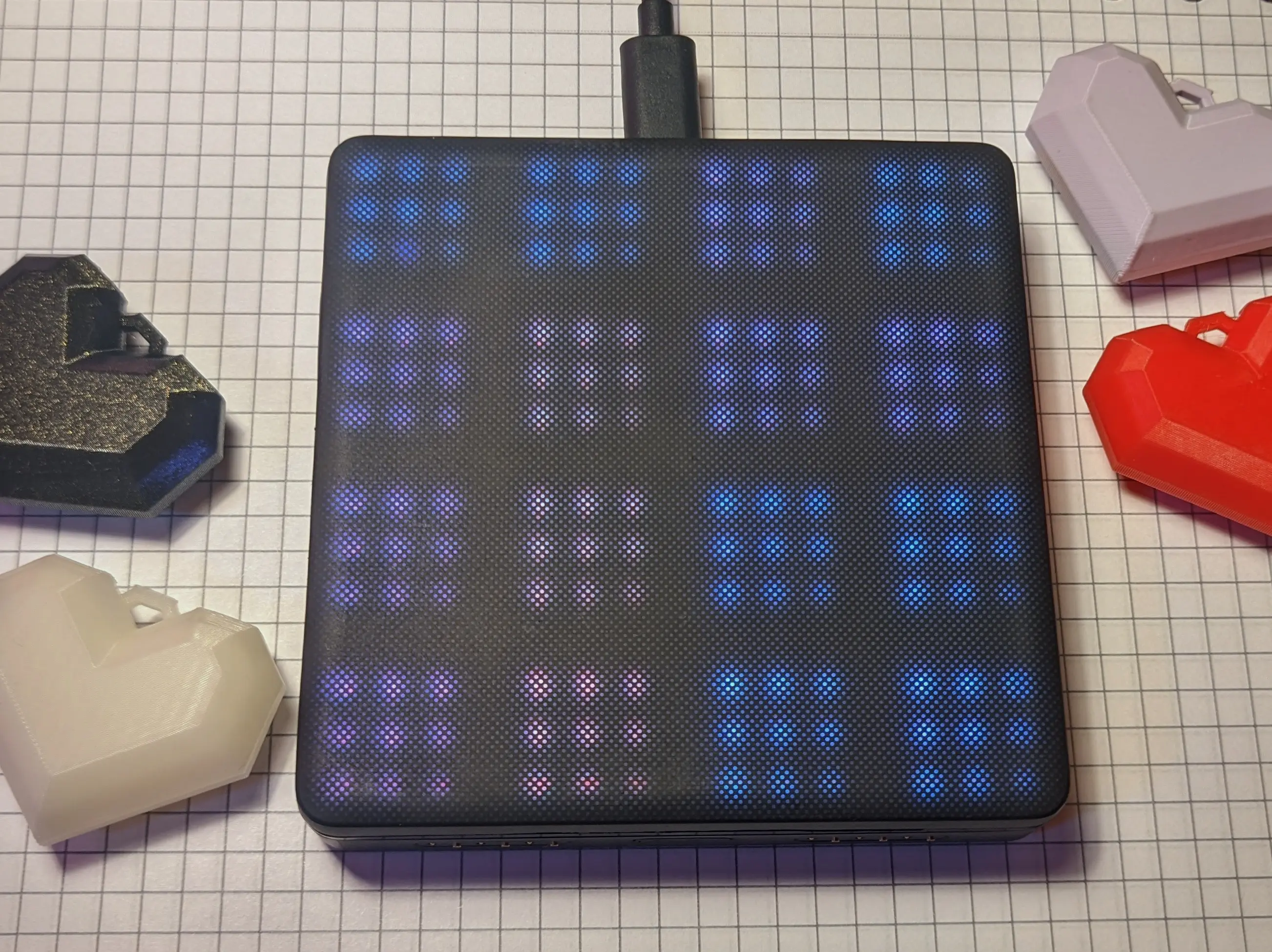 A larger block placed on a notebook. The block has rounded corners and appears more polished. Multiple groups of nine LEDs can be seen shining through the top silicone layer. A cable is running from the block to the top of the frame.
The notebook paper has been decorated with 3D-printed hearts.