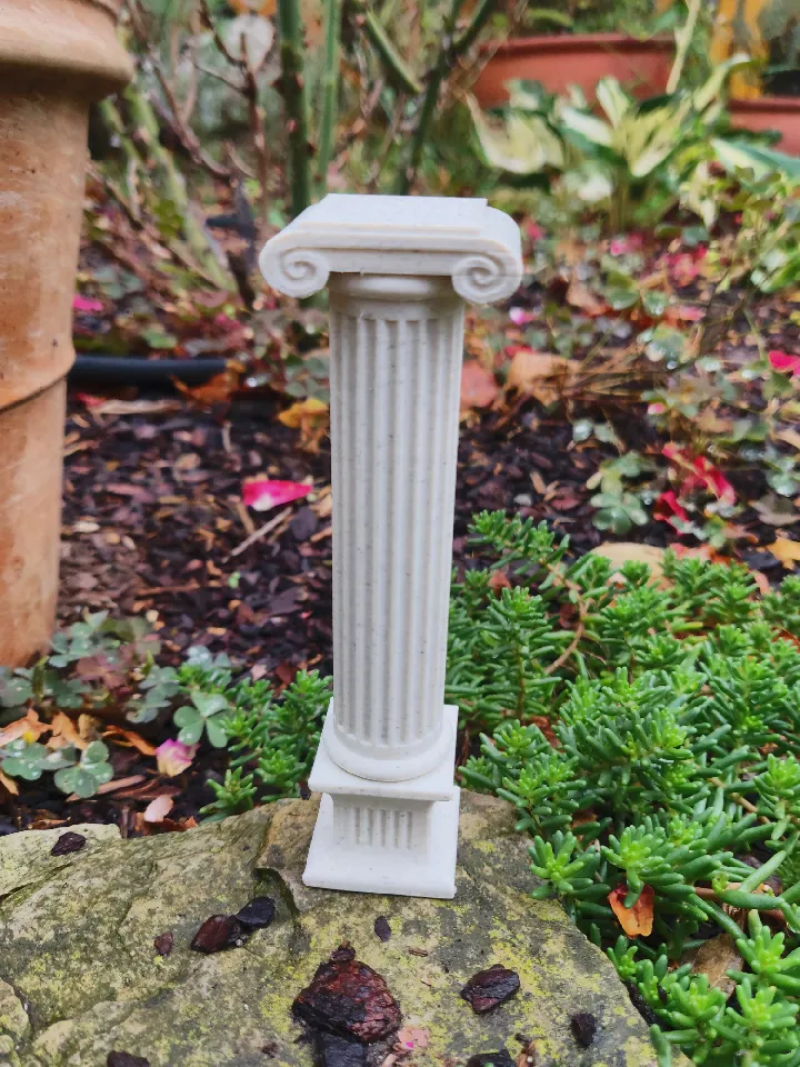 3D printed column standing on a small rock between plants. It has a square base, which transitions into a rounded shaft. It has a dual spiral on top