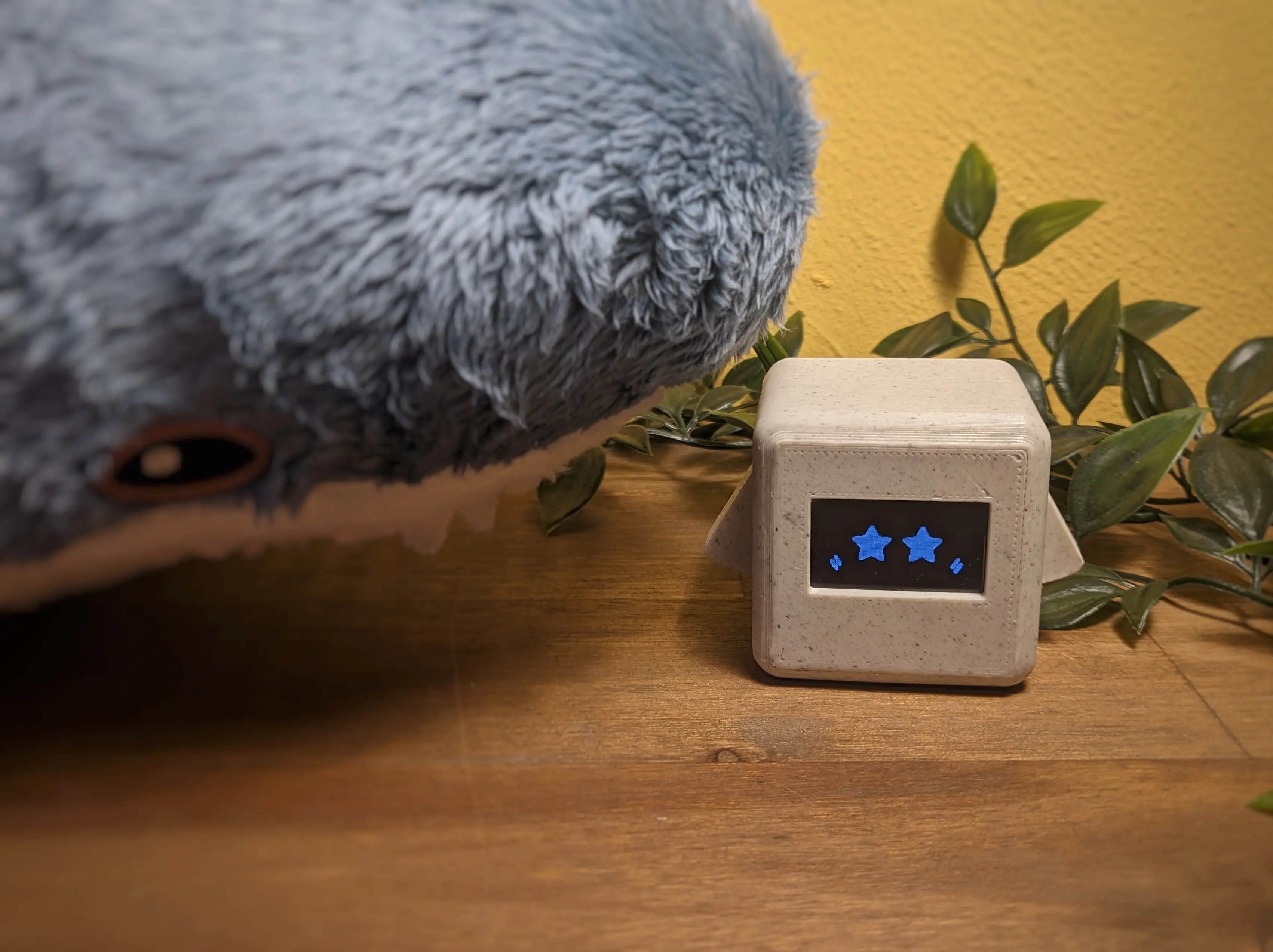 Precious Block with star eyes and blush shown on the display. The block is sitting in front of a plastic plant and next to a small Blahaj.