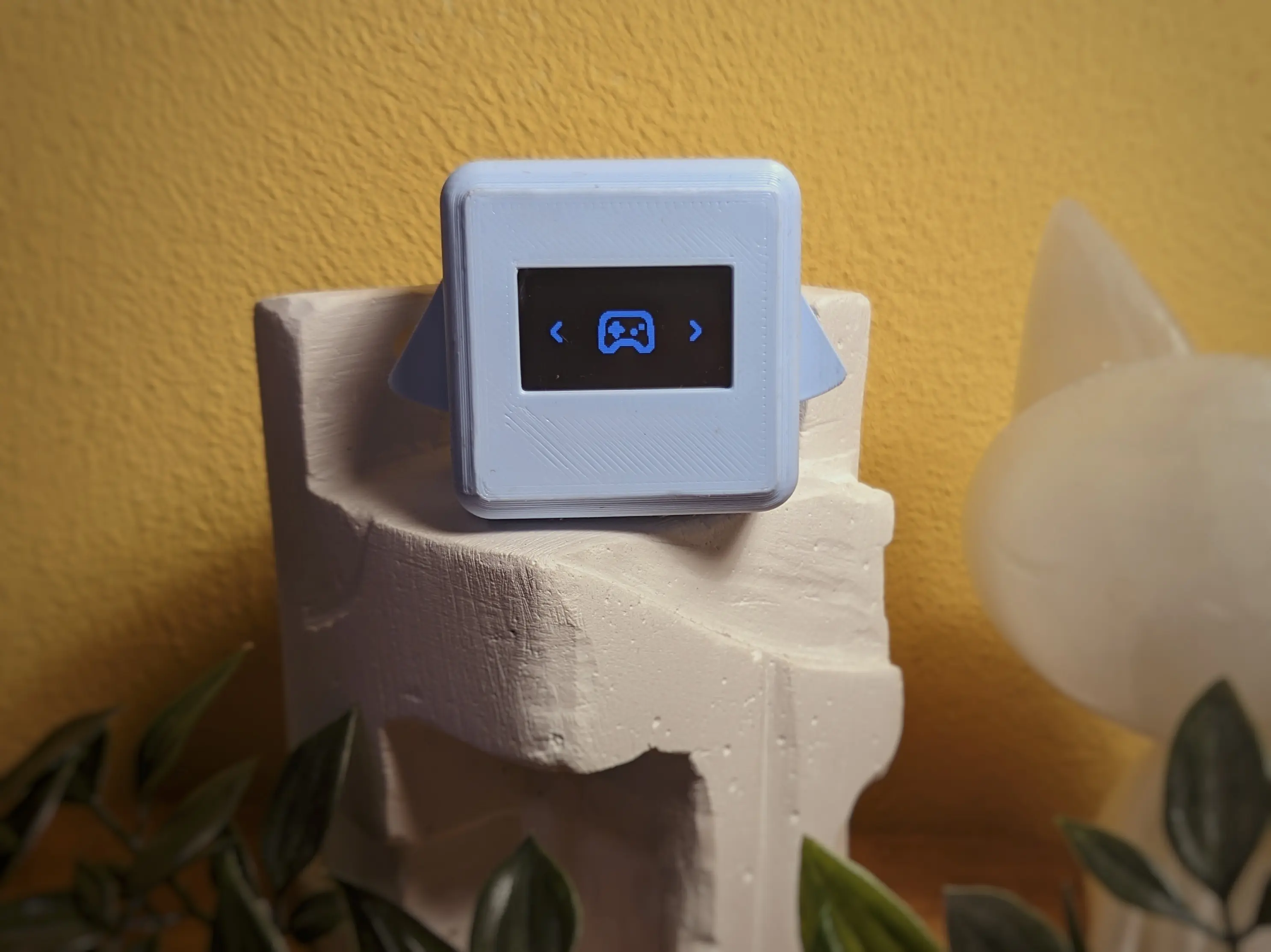 A blue Precious Block showing the menu UI. On both the left and right of the display are arrows. A game controller icon is centered on the display. The block has been placed on a white pedestal next to a small cat statue.