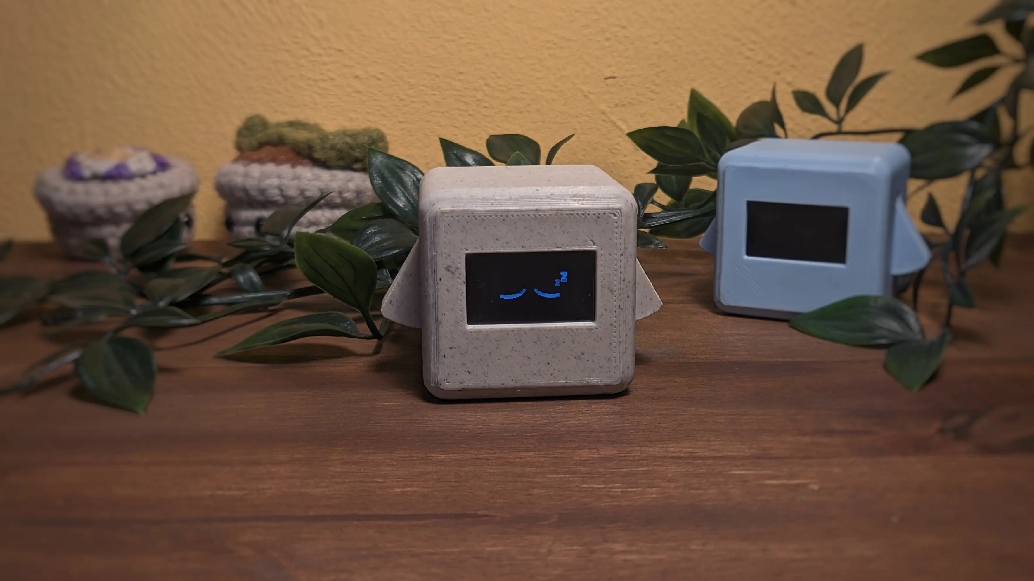 A Precious Block showing closed eyes and z symbols on its screen. In the background is a second blue block with its screen off. Next to it are other desk decorations, like crochet flowerpots and a plastic plant.