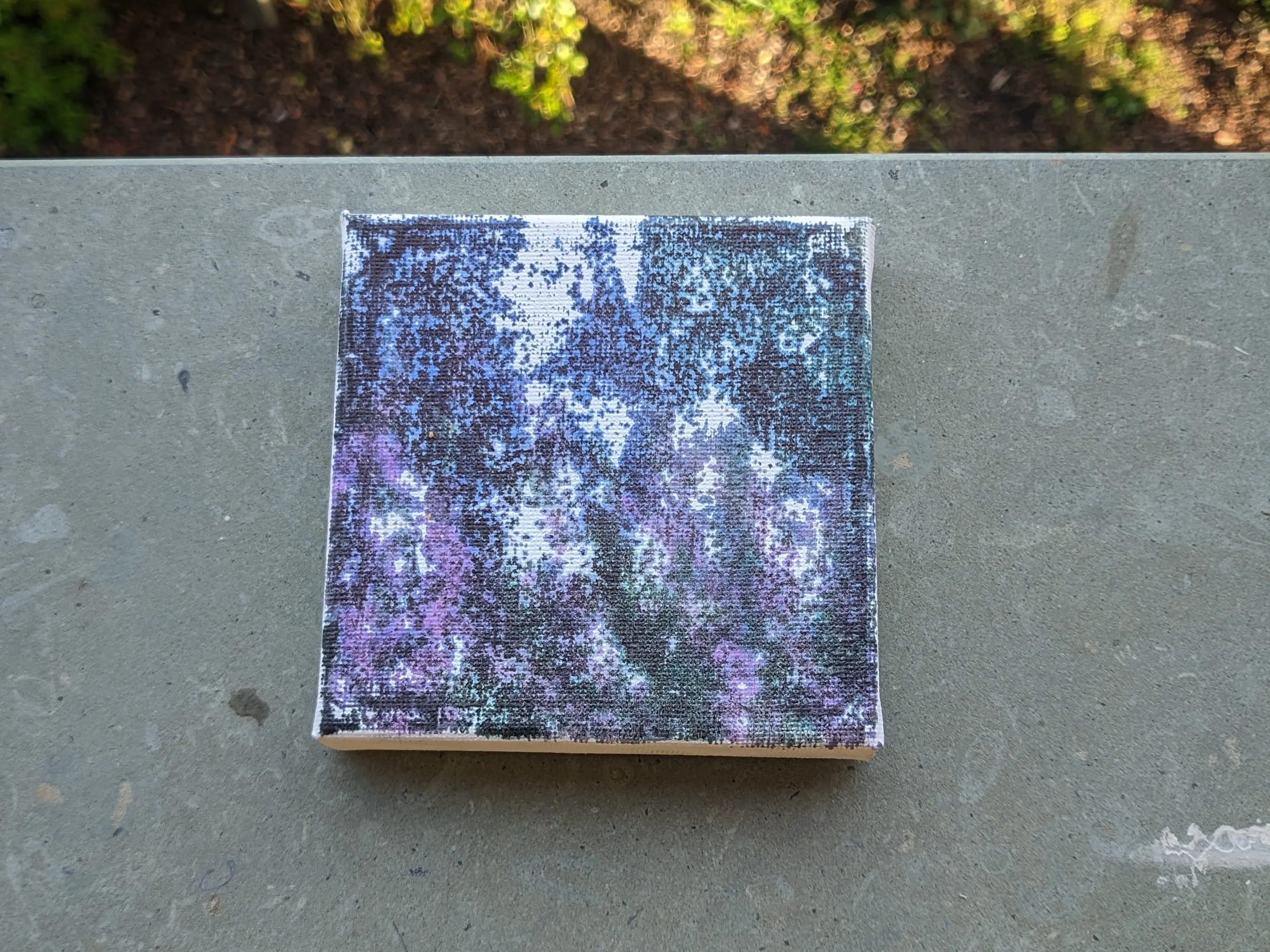 Top-down view of a small square canvas placed on a stone plate with plants in the background.
From a distance you can make out that the image transferred onto it is the same photo of a tree with purple flowers as above.
Up close, it becomes obvious that a lot of areas don&rsquo;t have ink on them at all, especially the brighter parts and the deep areas of the canvas.
The areas with enough ink also appear blurry.