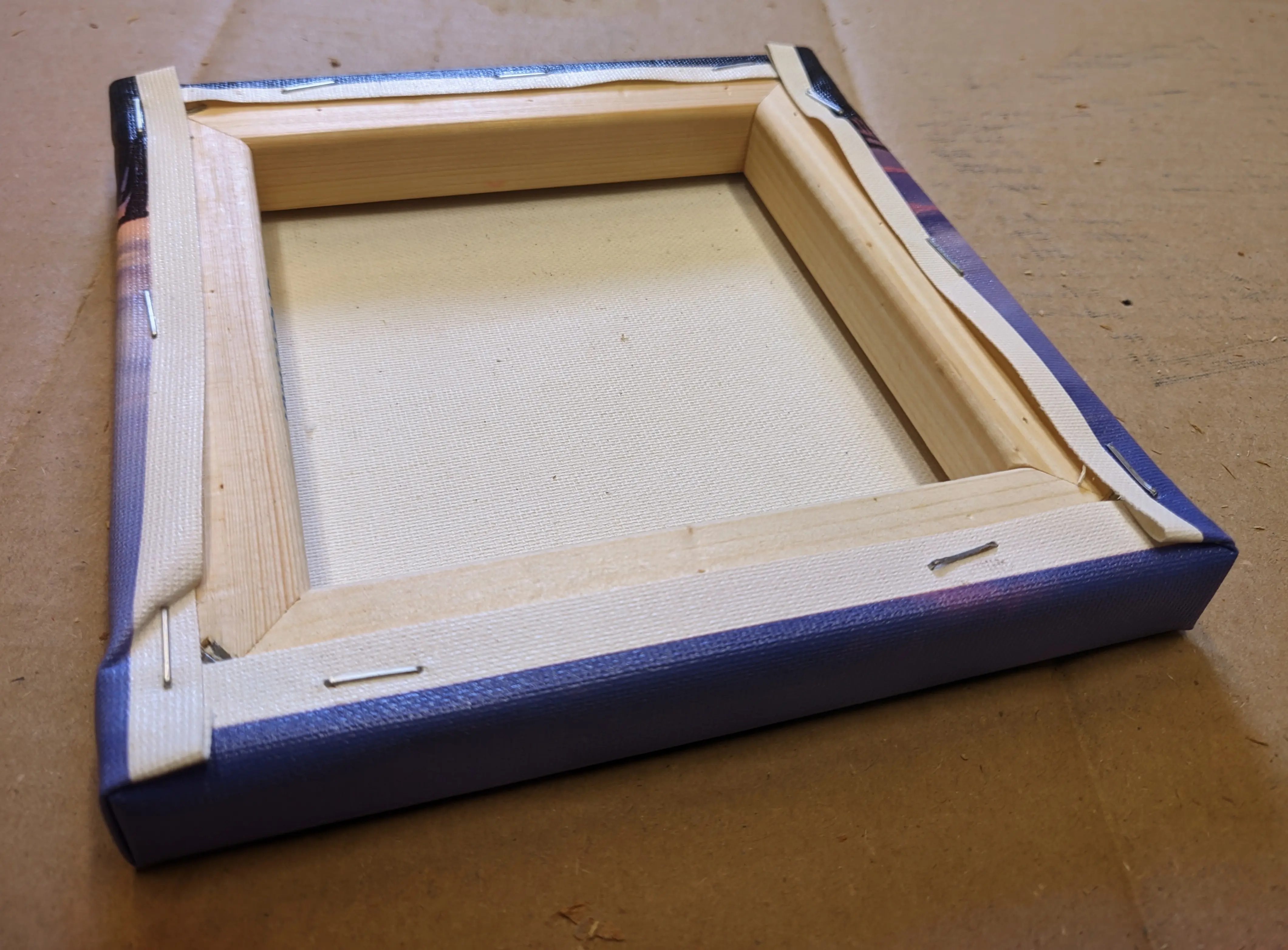The finished canvas lying with its back facing up on the cardboard sheet.
Multiple staples running along all edges are pinning the canvas sheet to the frame.
Most of the staples are placed directly on the transition area from white canvas to printed canvas.