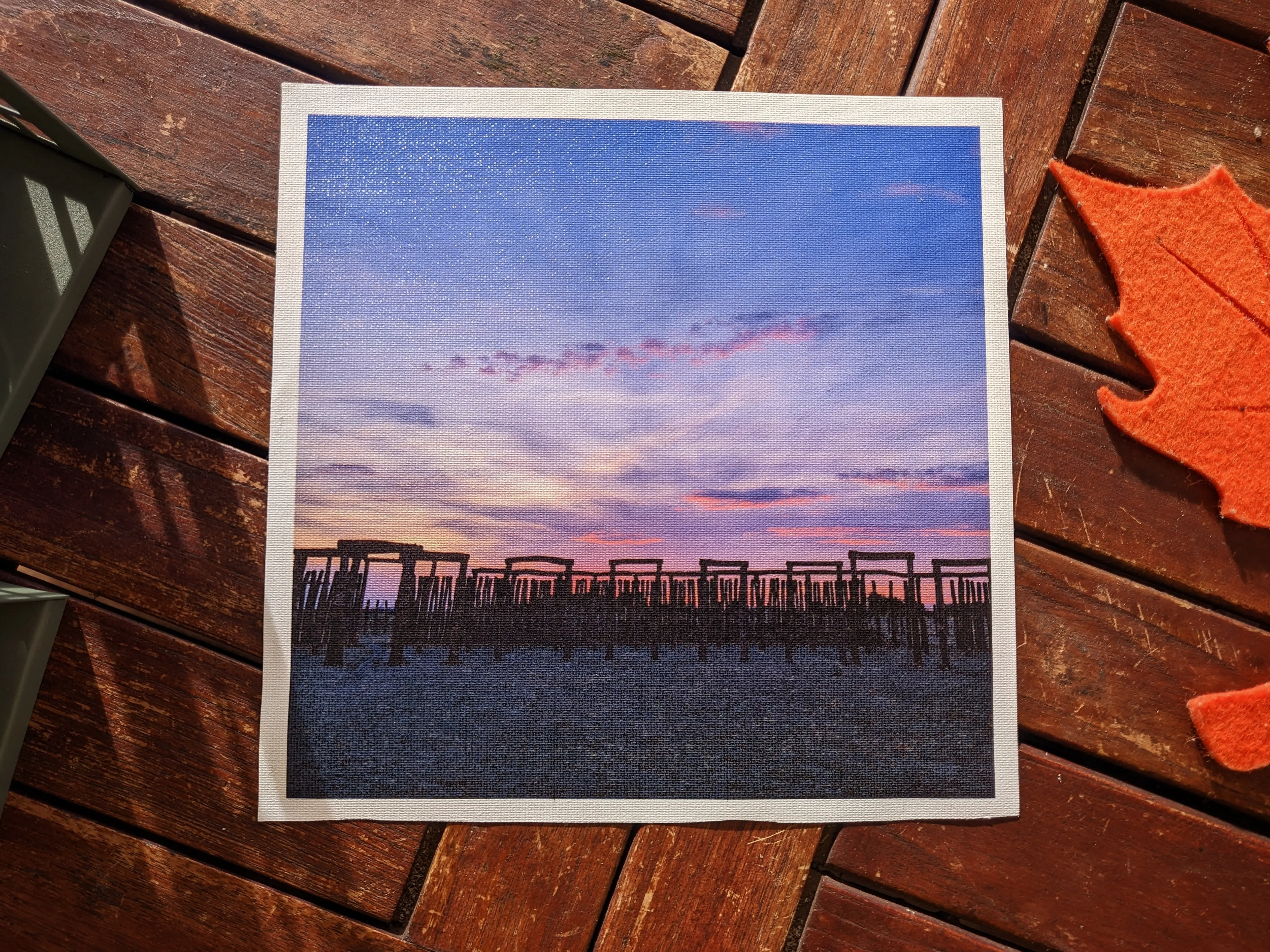 Square cutout of a canvas sheet placed on a wooden desk.
The sheet has a printed image of a wooden structure at dusk on it, with white margin on all four sides.