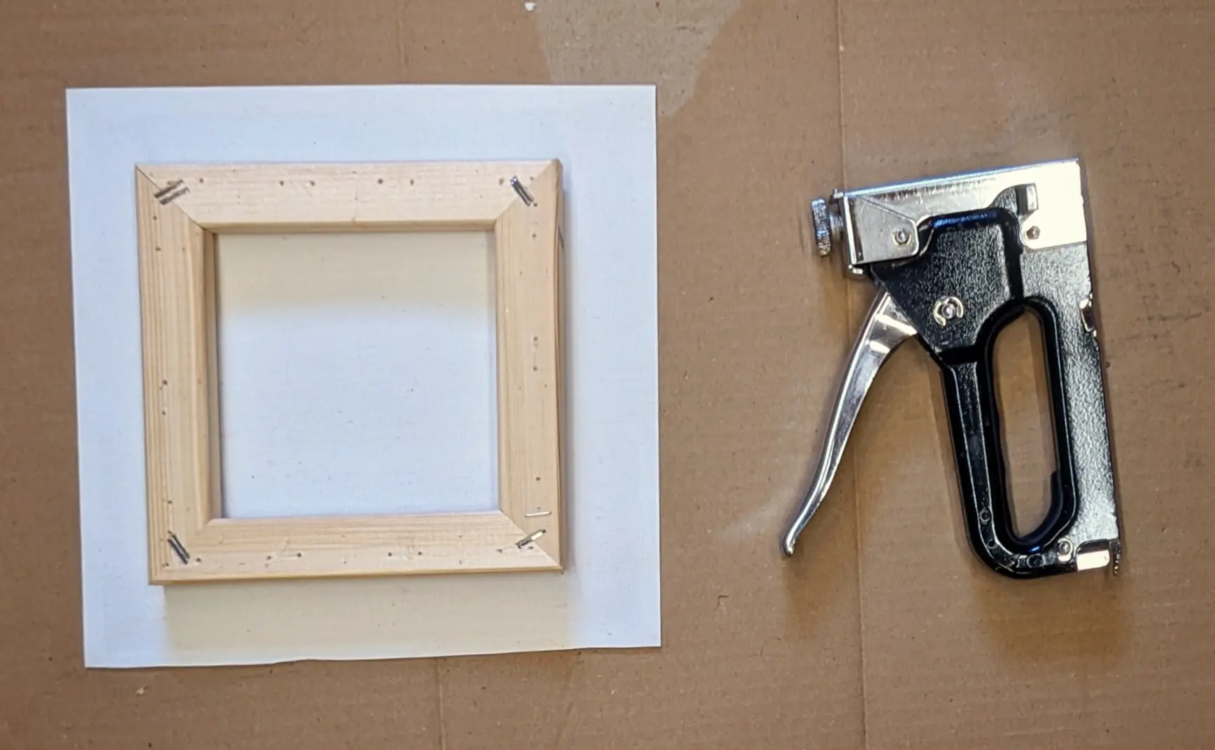 Top-down view of a cardboard sheet.
On the left, the canvas sheet has been placed below the canvas frame with the back facing up. The canvas sheet is larger than the frame and extends below it in all direction.
On the right, a light duty staple gun has been placed.