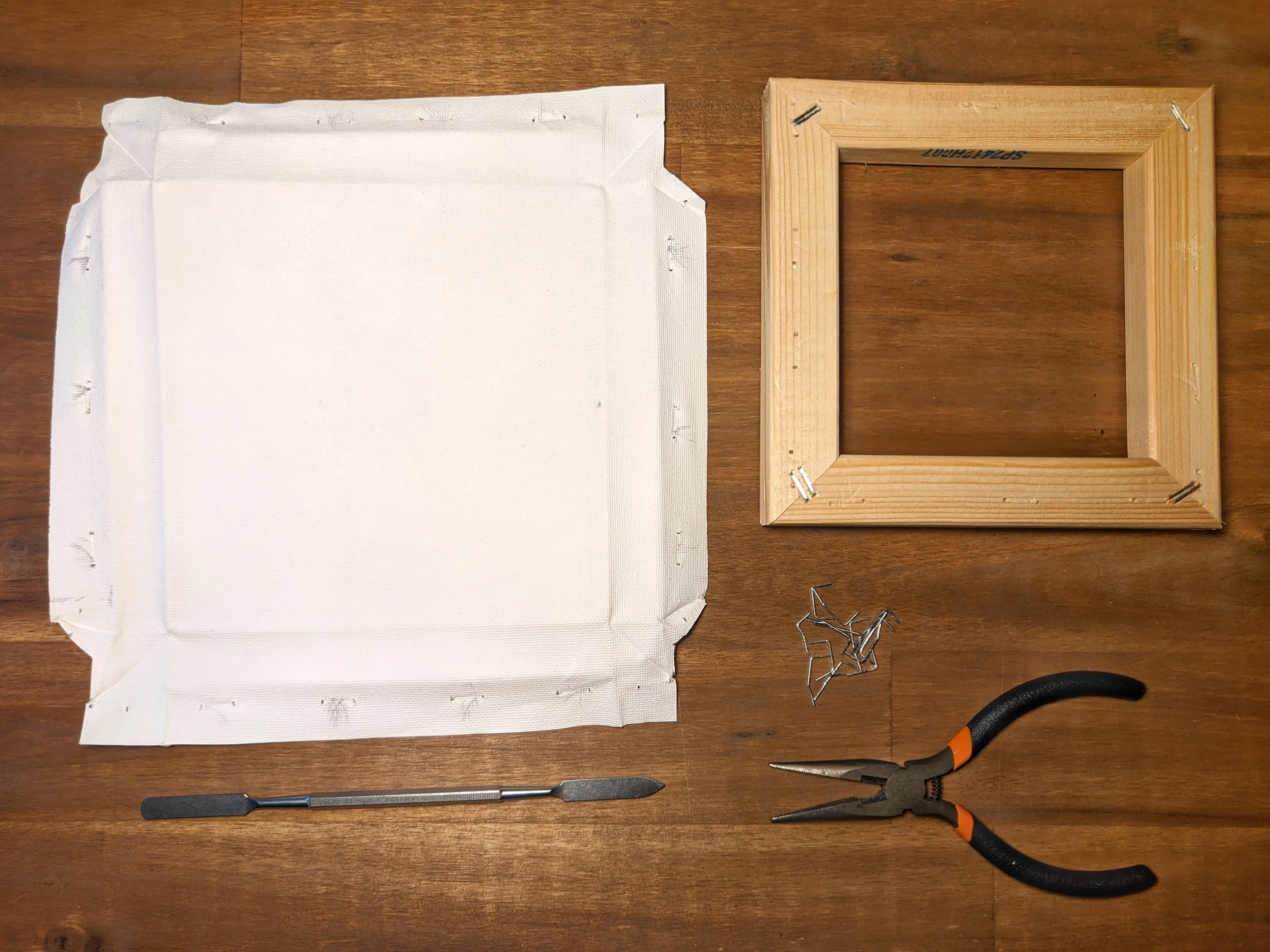A disassembled canvas frame placed on a wooden desk.
The canvas fabric has been folded flat and is sitting next to the wooden frame.
A metal wedge, a pair of pliers, and the removed staples have been placed beneath the canvas sheet.