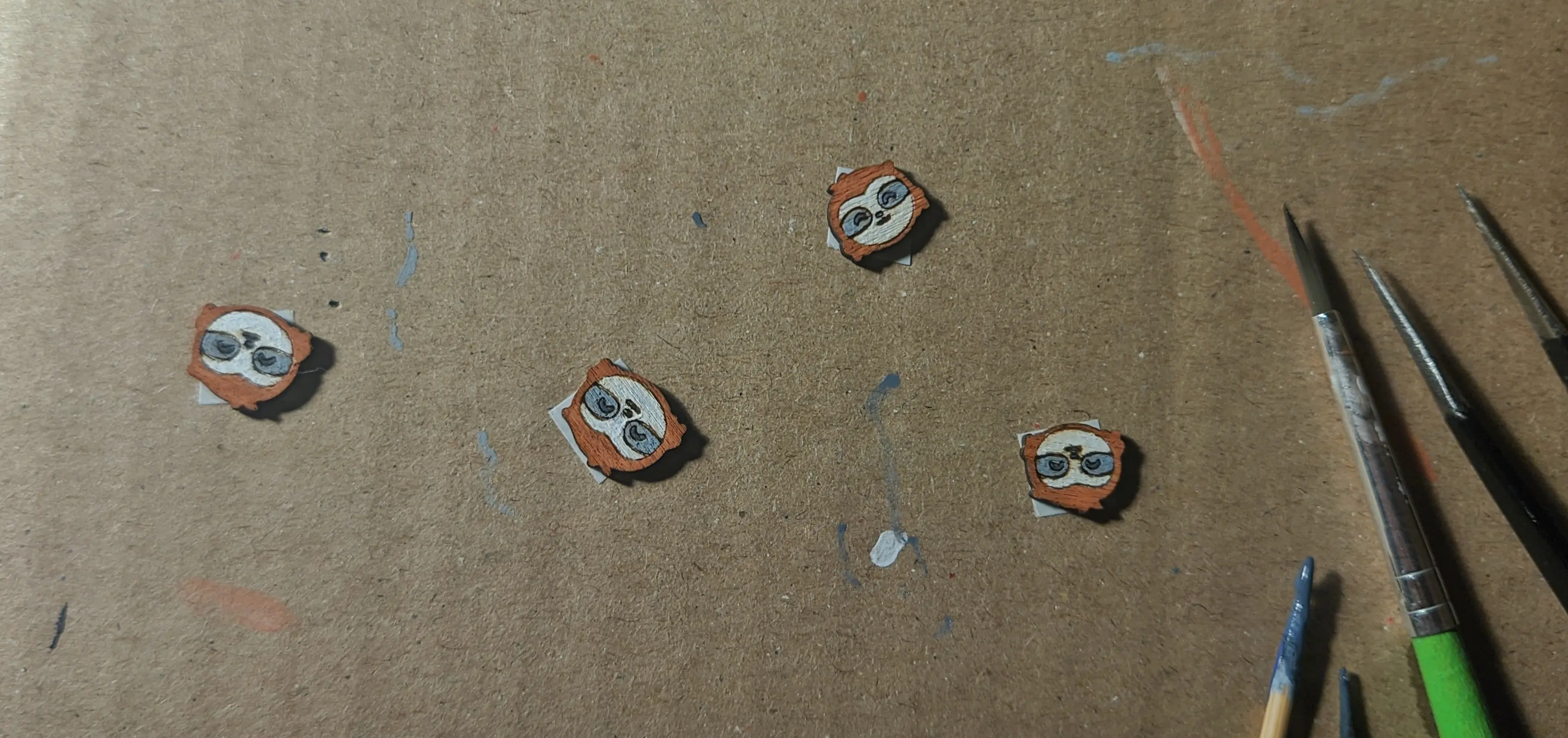 All four wooden sloths stuck to a piece of cardboard and fully painted in brown, white, gray, and black. A toothpick, a paintbrush, and a pair of tweezers are placed next to it. The cardboard has some paint marks on it.