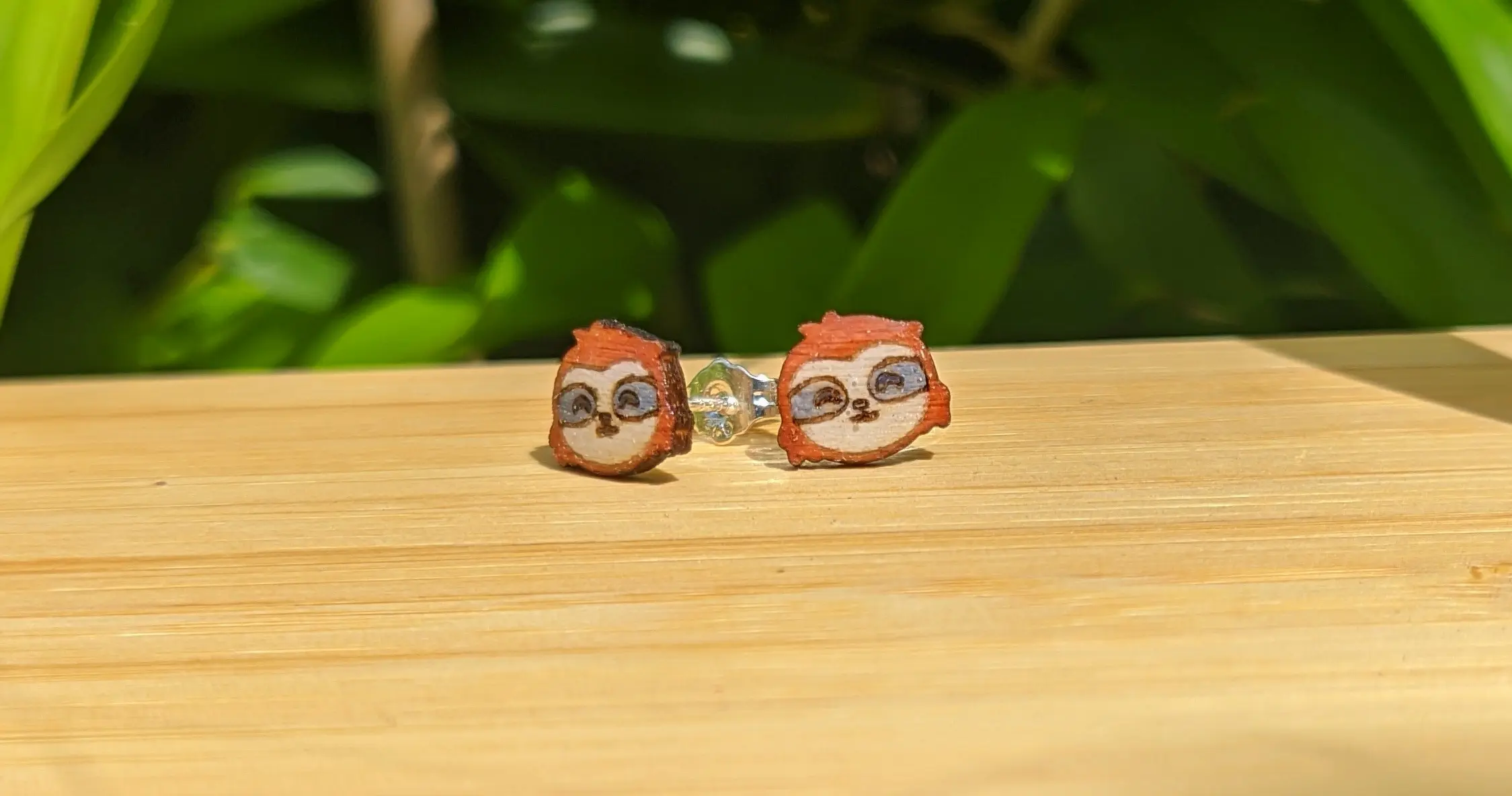 Finished earrings sitting on a piece of wood. The camera is directly facing the sloth face, revealing the color and engraving. One of the earrings is placed at a slight angle, revealing the earring post behind.