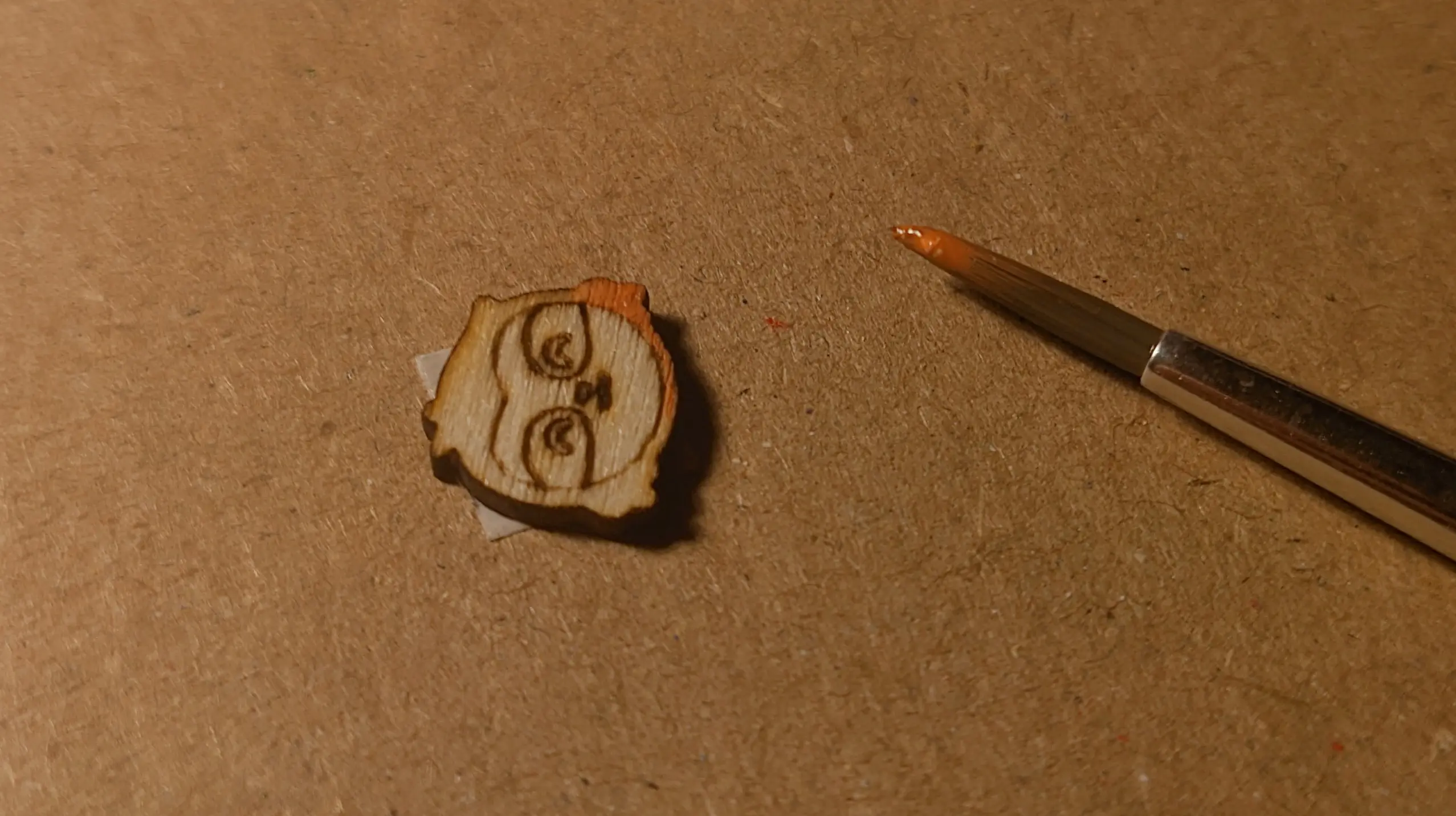 A single wooden sloth stuck to a piece of cardboard. One side of its face has already been painted orange-brown. A small paintbrush, with color on it, is lying next to it.