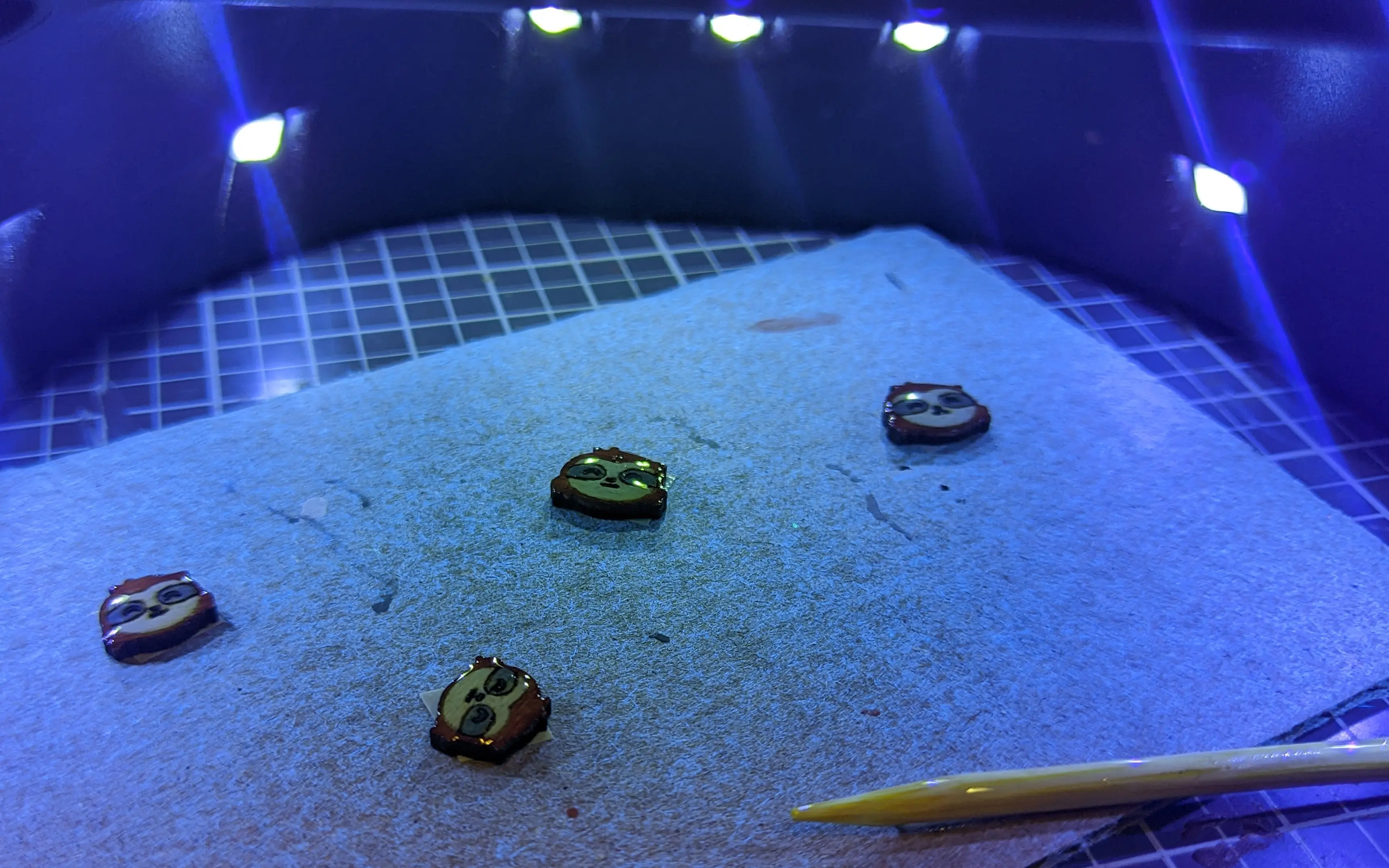 Piece of cardboard with the wooden sloths pieces attached lit by a UV light. Every piece is covered in transparent resin, which creates a slight curve on the objects.