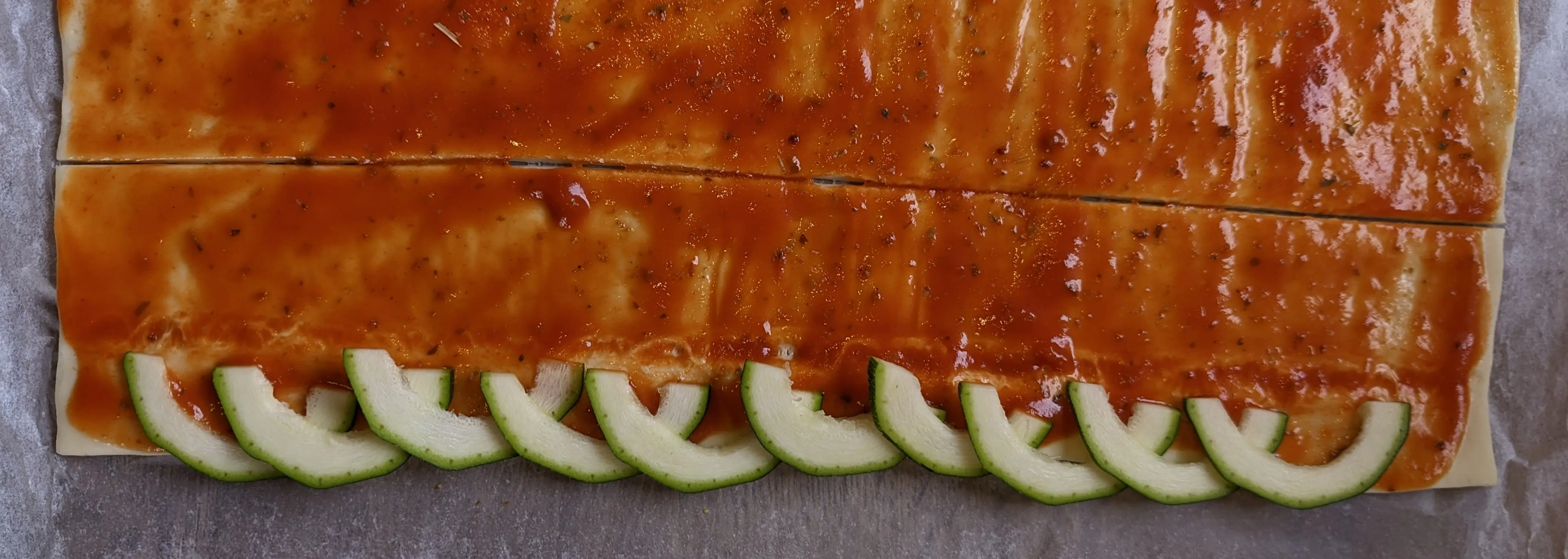 The rainbow-shaped Zucchini slices are laid out along the side of the dough, slightly overlapping the edge. The dough has been cut about two slices wide from the edge.