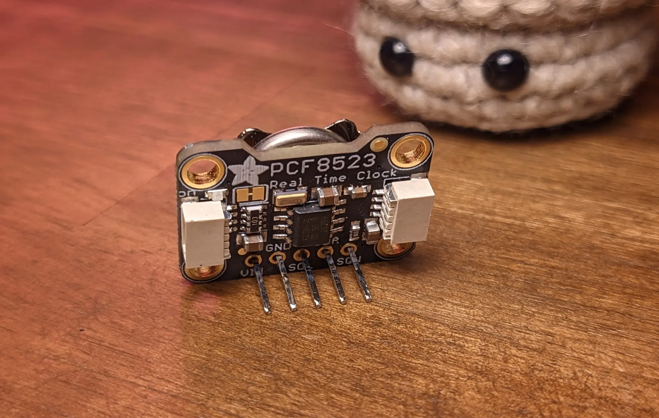 Close-up of a circuit board with the text PCF8523 printed on it. In the background the eyes of the tiny crochet flowerpot can be made out.