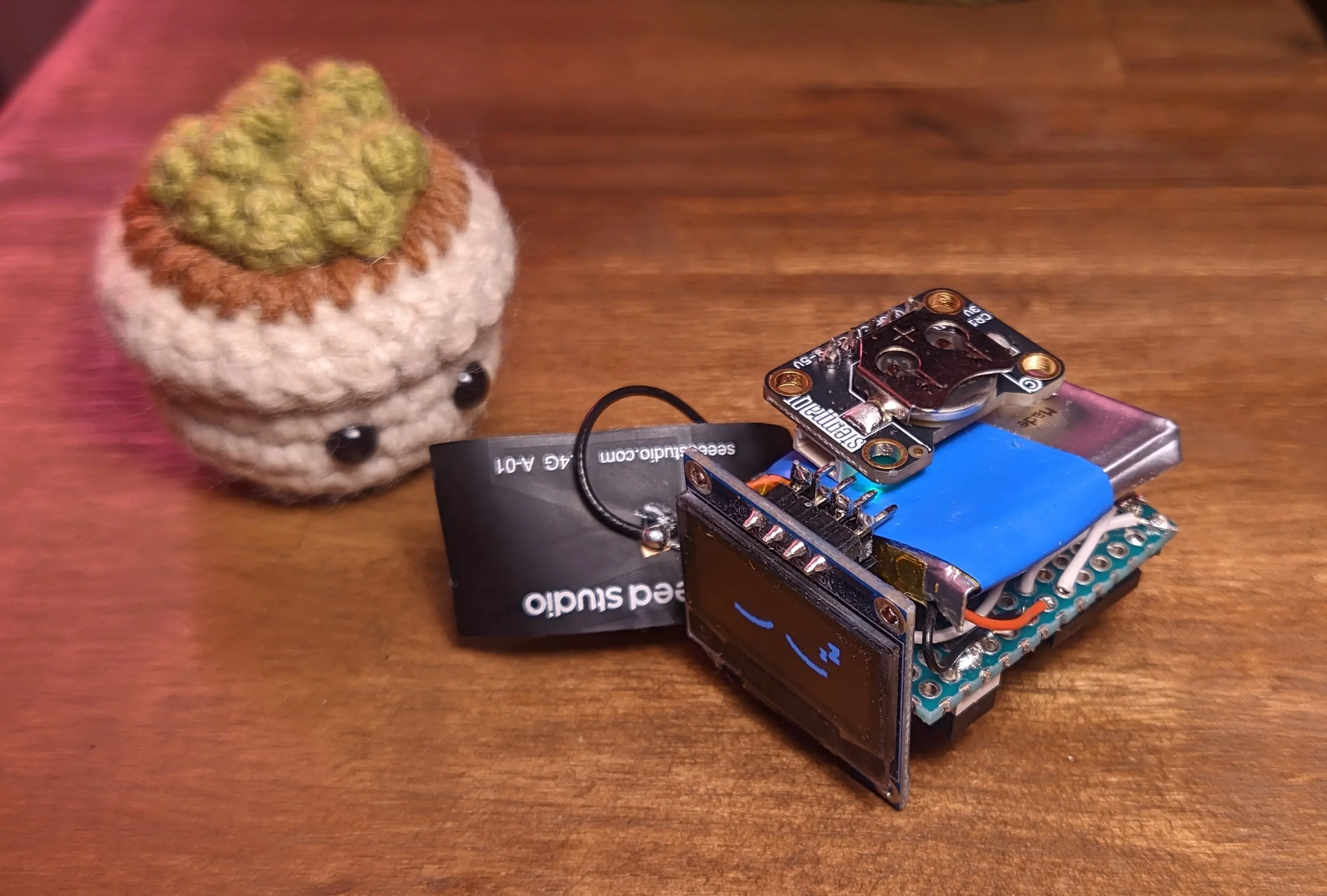 The tiny crochet flowerpot looking at a stacked protoboard with a display on the front and a battery and the clock module on top. The screen is turned on and displaying the same sleeping face as earlier.