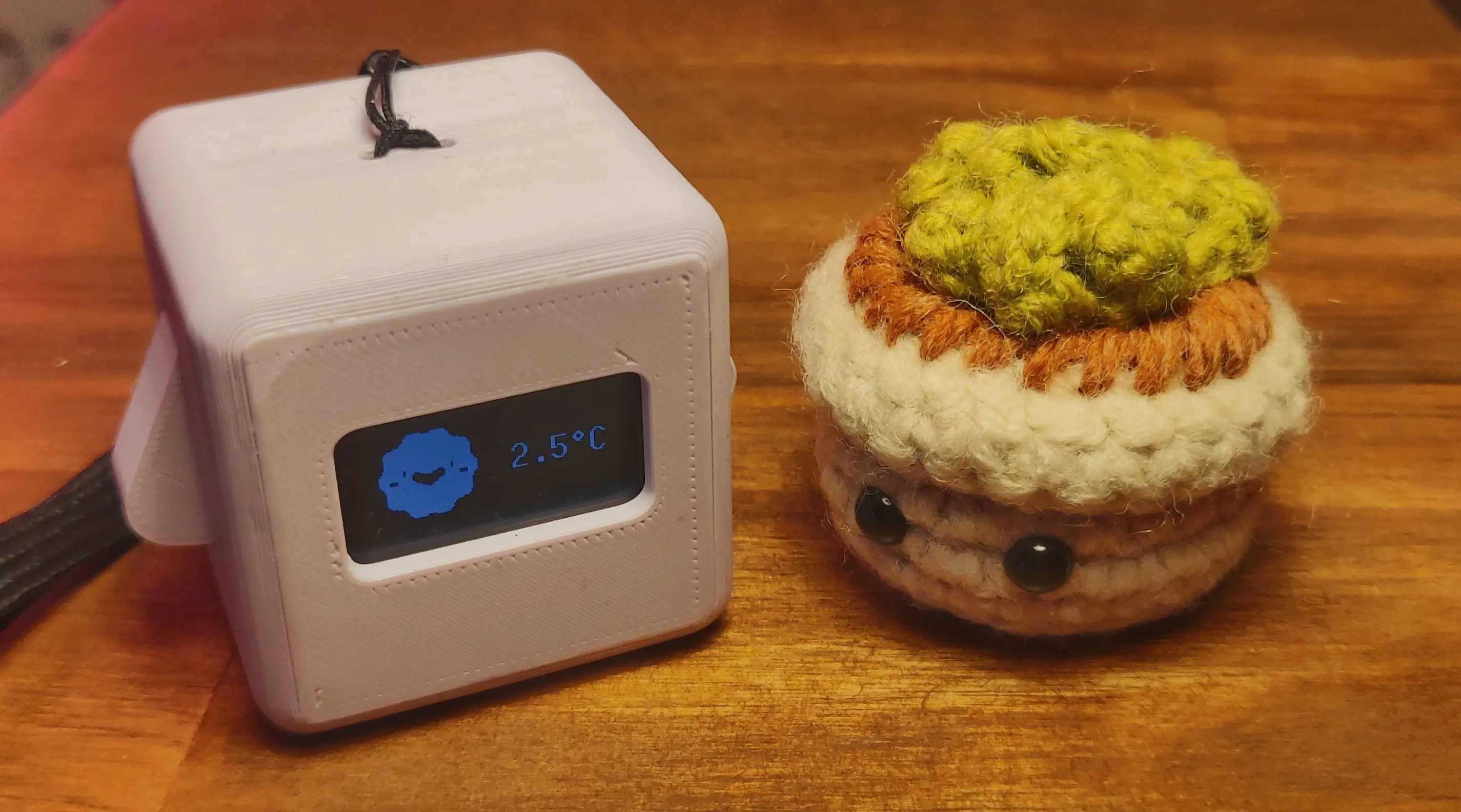 A Precious Block sitting on the left of the tiny crochet flowerpot.
Its display is showing a smiling sun next to a temperature indicator reading 2.5°C.