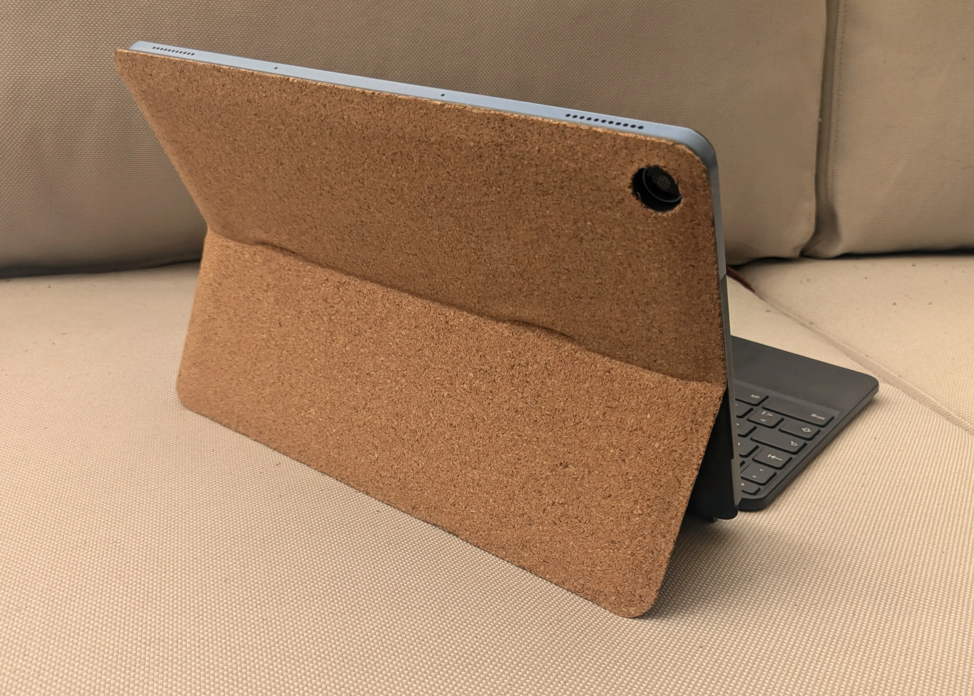 Tablet with a fine-grain cork case stood up on a sofa. The lower half of the case has been folded out to support the device at an angle. A keyboard is connected to the bottom of the device