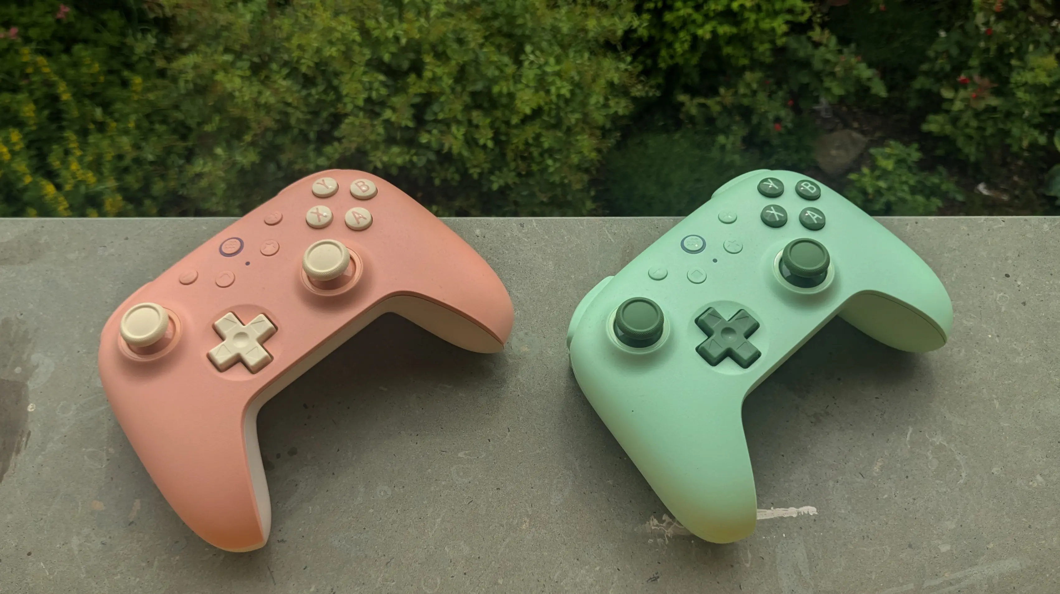 Two pastel-color game controllers placed on a stone window sill, with greenery showing below them.
