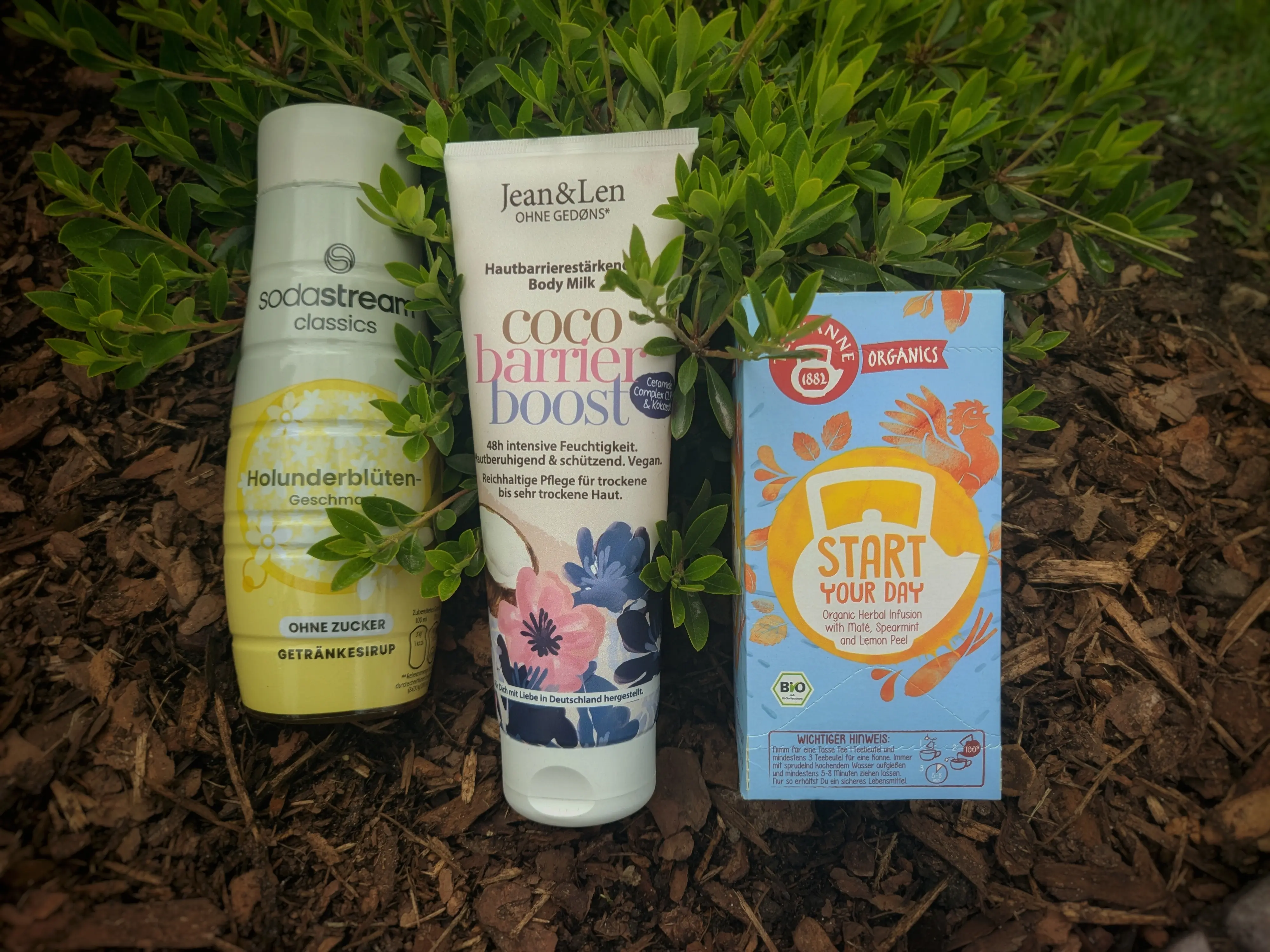 Three items resting against a rhododendron bush, which has been planted on mulch.
The left most item is a cylindrical bottle, which bulges out towards the bottom. It is labeled Sodastream classics elderflower (in German) with drawn elderflowers.
Next to it is a large tube labeled Jean & Len coco barrier boost body milk, with illustrations of flowers and an opened coconut on it.
The right most item is a cardboard tea box, labeled Teekanne organics start your day. The box is a light shade of blue with orange graphics on it.