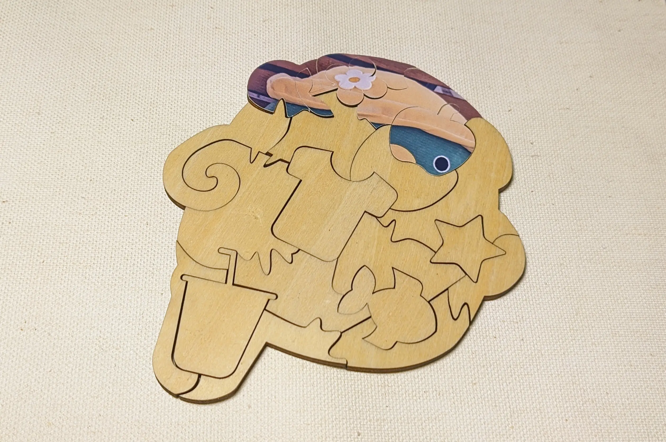 Assembled blob-shaped wooden puzzle on a white backdrop.
A couple of the pieces have stickers with a printed graphic applied to them, but most of them are still bare.
You can make out the shapes of the bare pieces fairly easily. There is a T-shirt, a star, a fish, and a bubble tea. The other pieces are random shapes.