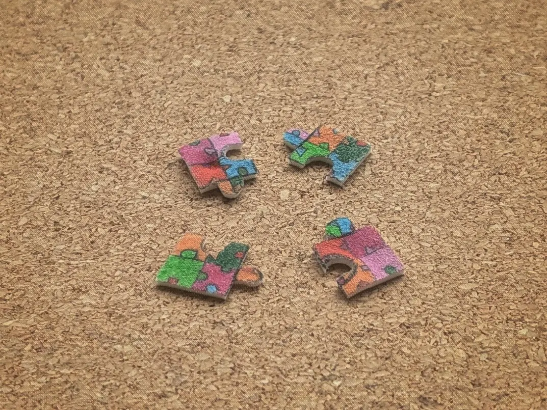 Four small, colourful puzzle pieces placed on a wooden surface. They each have multiple tiny puzzle pieces drawn onto them.
The pieces throw irregular shadows onto the surface they have been placed on, revealing that they are not entirely flat.