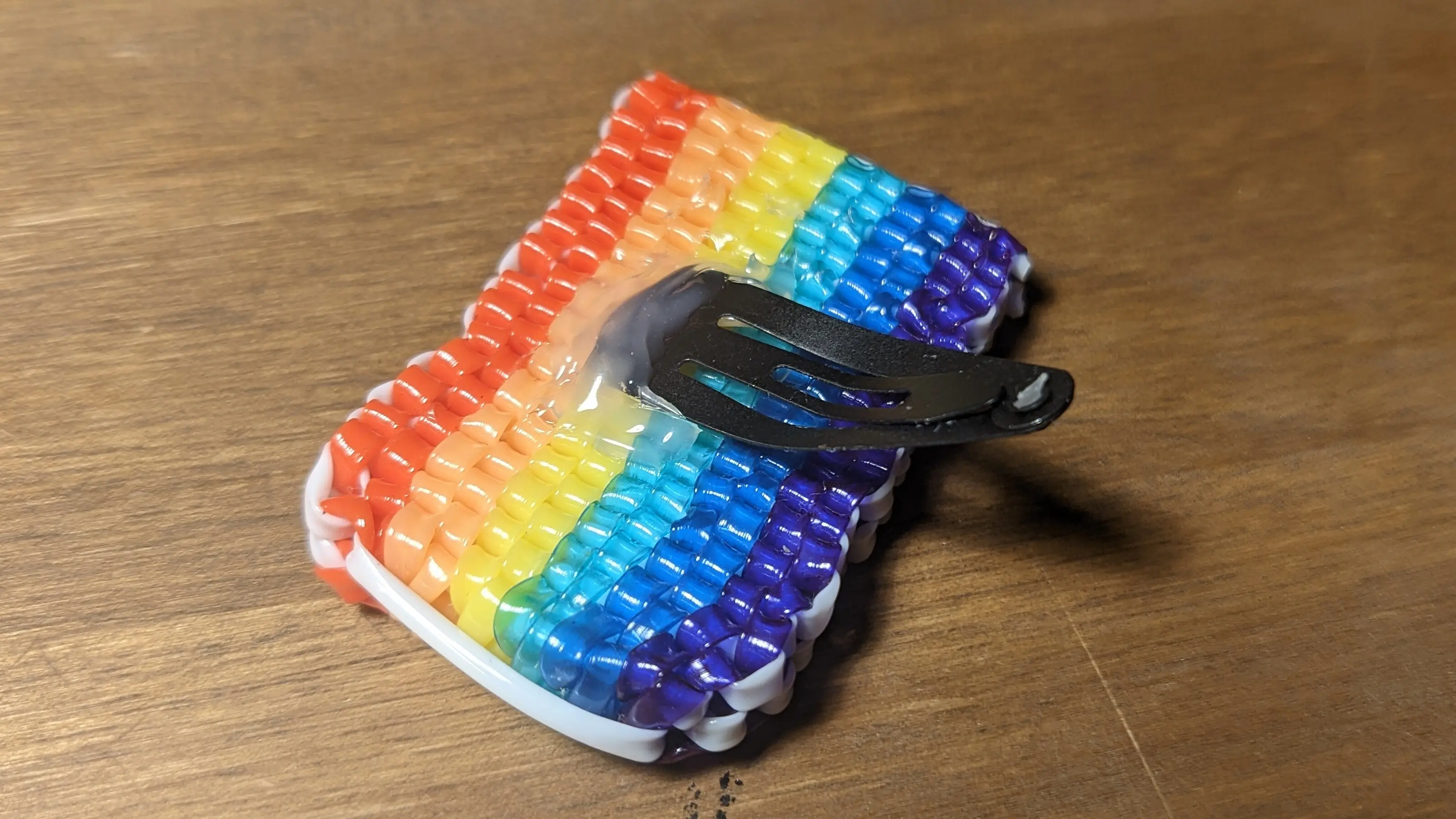 Back of the rainbow flag, with a hairpin hot-glued to it. The hairpin has been glued to the center of the flag and extends from the shorter side.