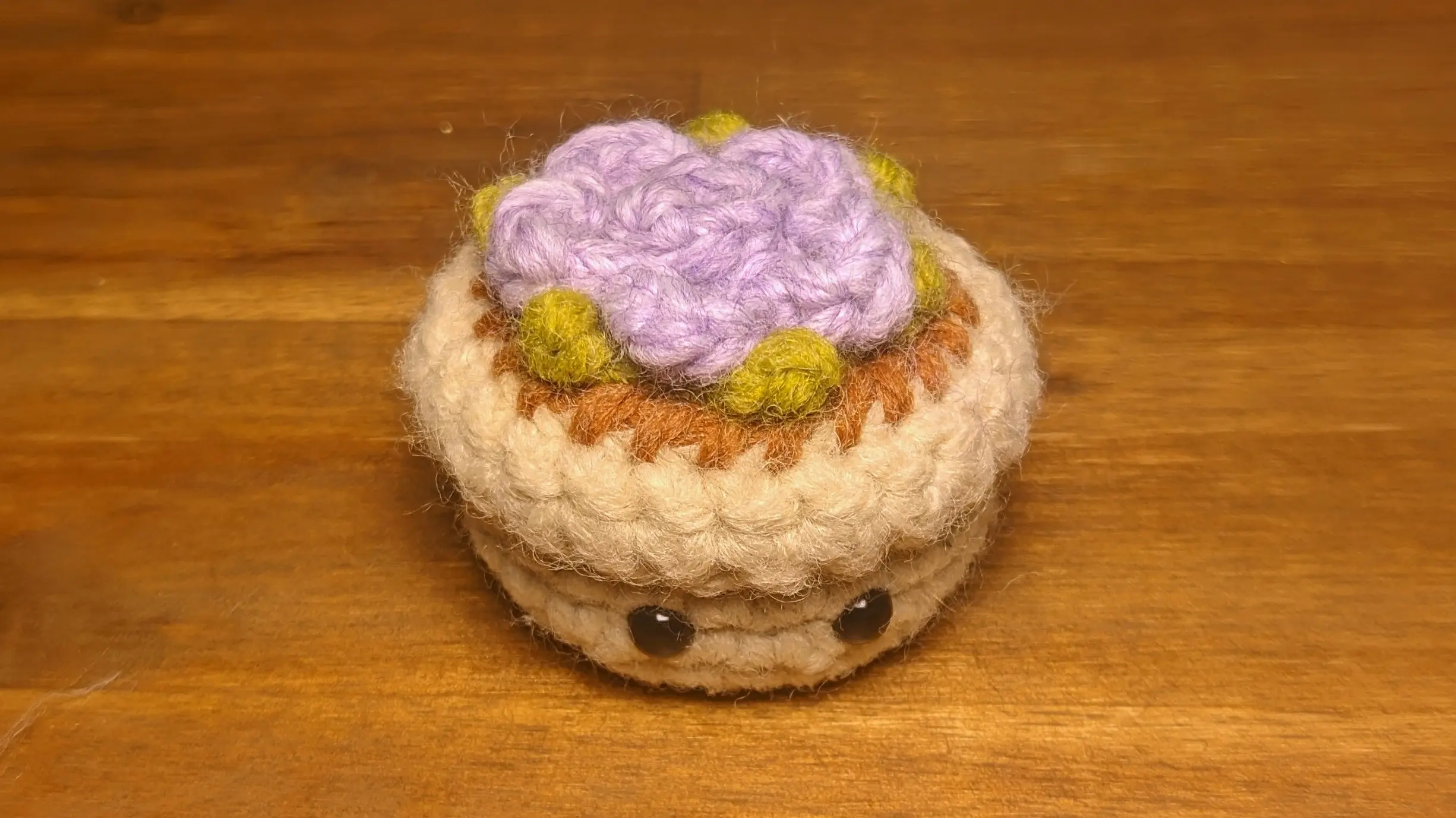 Close-up of the tiny crochet flowerpot with a lavender-colored flower on top. The tips of the green leaves extend from underneath the flower petals.
The flower itself is rose-like in appearance and spirals inwards.