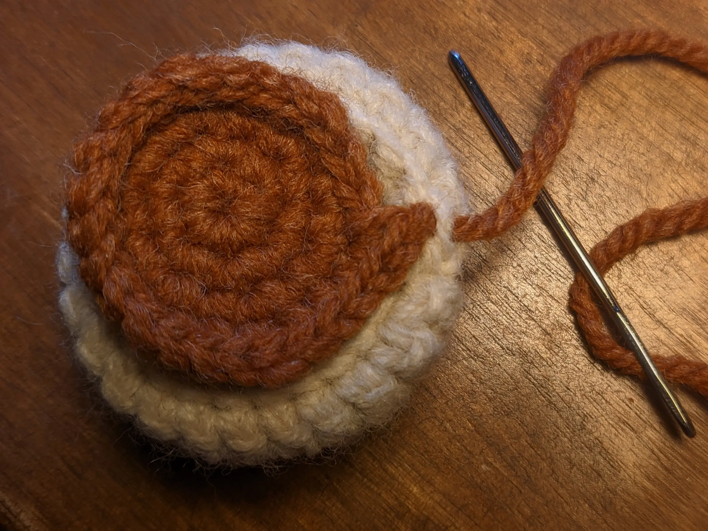 The small, bumpy end section of the brown crochet disk aligned with the end of the round of the base pot.
The remaining yarn of the brown disk has been pulled through the previously unused inner loop of the main wall&rsquo;s top row upwards.