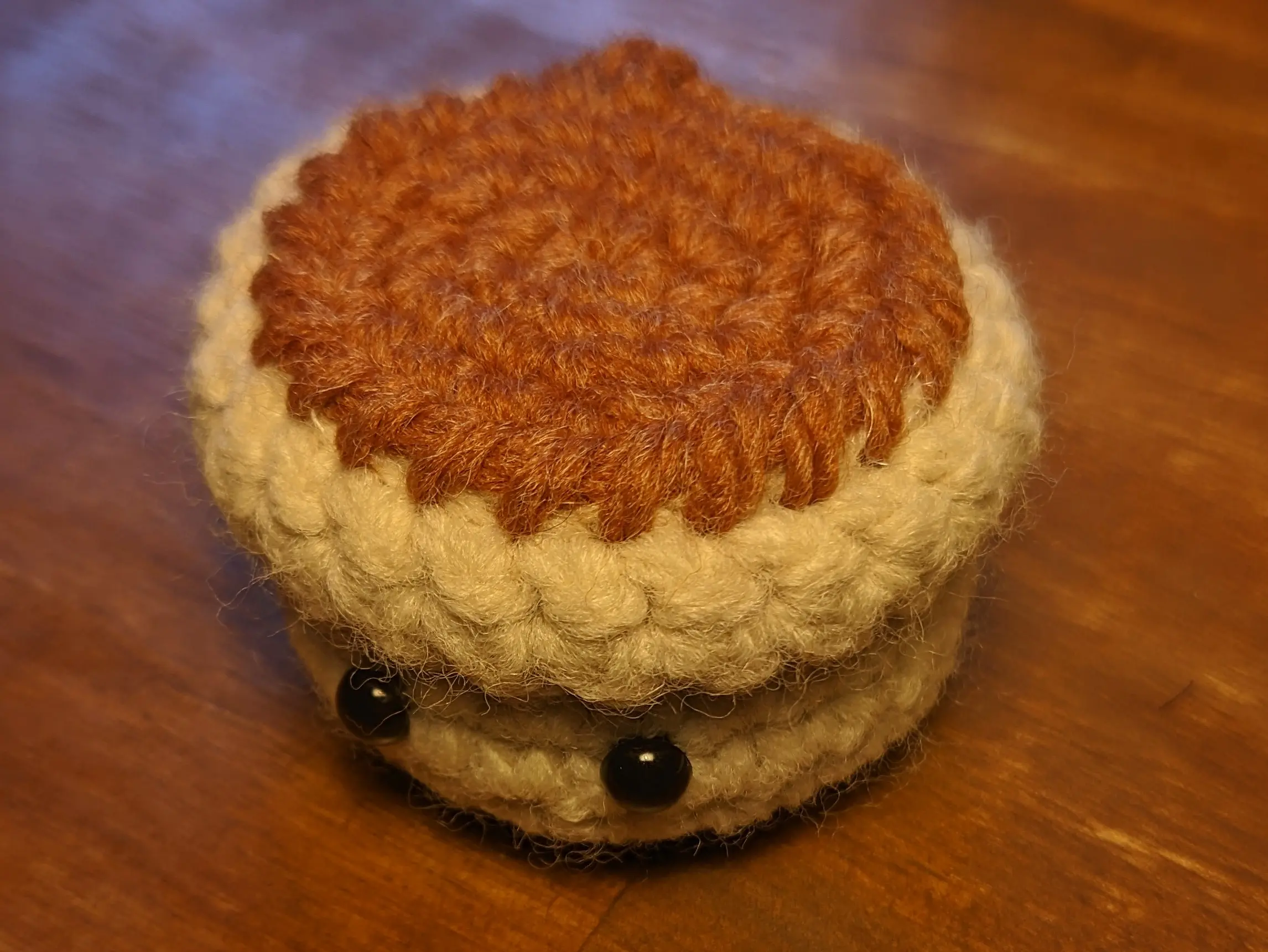 A small crochet pot placed on a wooden desk. The pot itself is a white/sand color with a brown topsoil sewn to it on the top, slightly extending from the top of the pot.
The pot has small black plastic eyes and is slightly turned from the camera. The glow of a lamp is reflected in the eyes from above.
Typical crochet stitch patterns are visible on both the outside of the pot and the top layer.