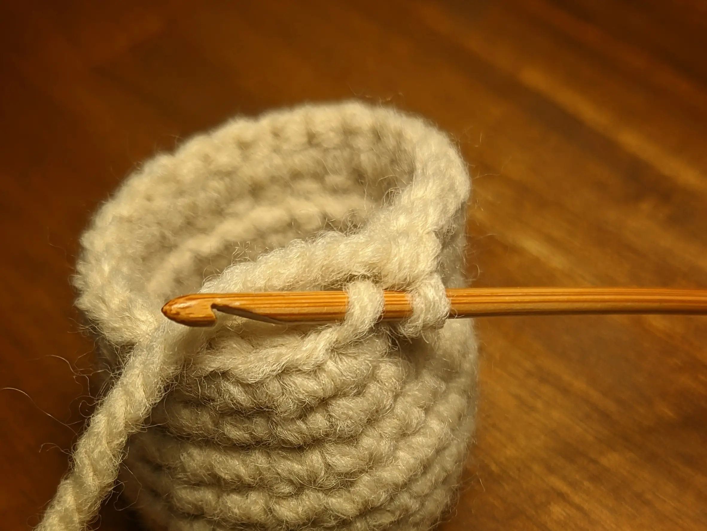 The wooden crochet hook has been placed through the outer loop of the top cylinder wall stitch, running from bottom to top. The inner loop is hidden by the hook and the yarn.
